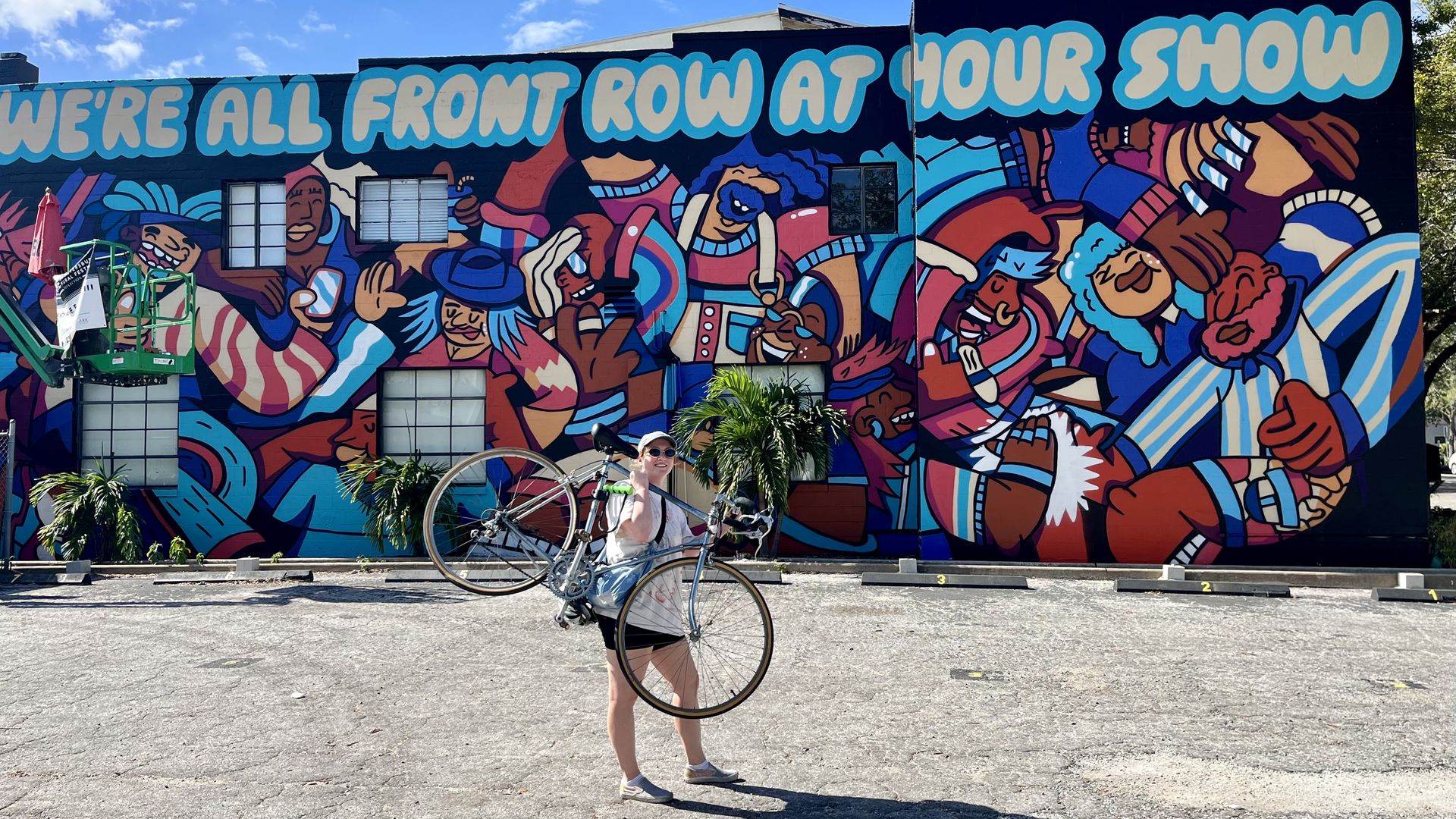A woman hoisting a road a bike with a light blue frame, smiling at the camera, in front of a colorful mural featuring people dancing that says "We're all front row at your show."
