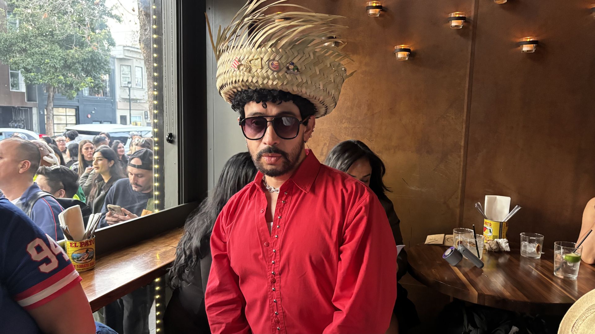 Man wearing a large woven straw hat with pins, dark sunglasses, a red button-up shirt, and a silver chain necklace, standing inside a dimly lit cafe with wooden tables and a brown wall with small lights.