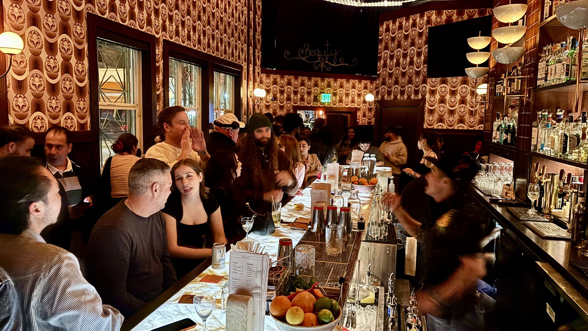 Crowded bar with patterned brown wallpaper, people sitting and chatting along a glowing marble countertop, bartender in motion, shelves with bottles and glasses on the right.
