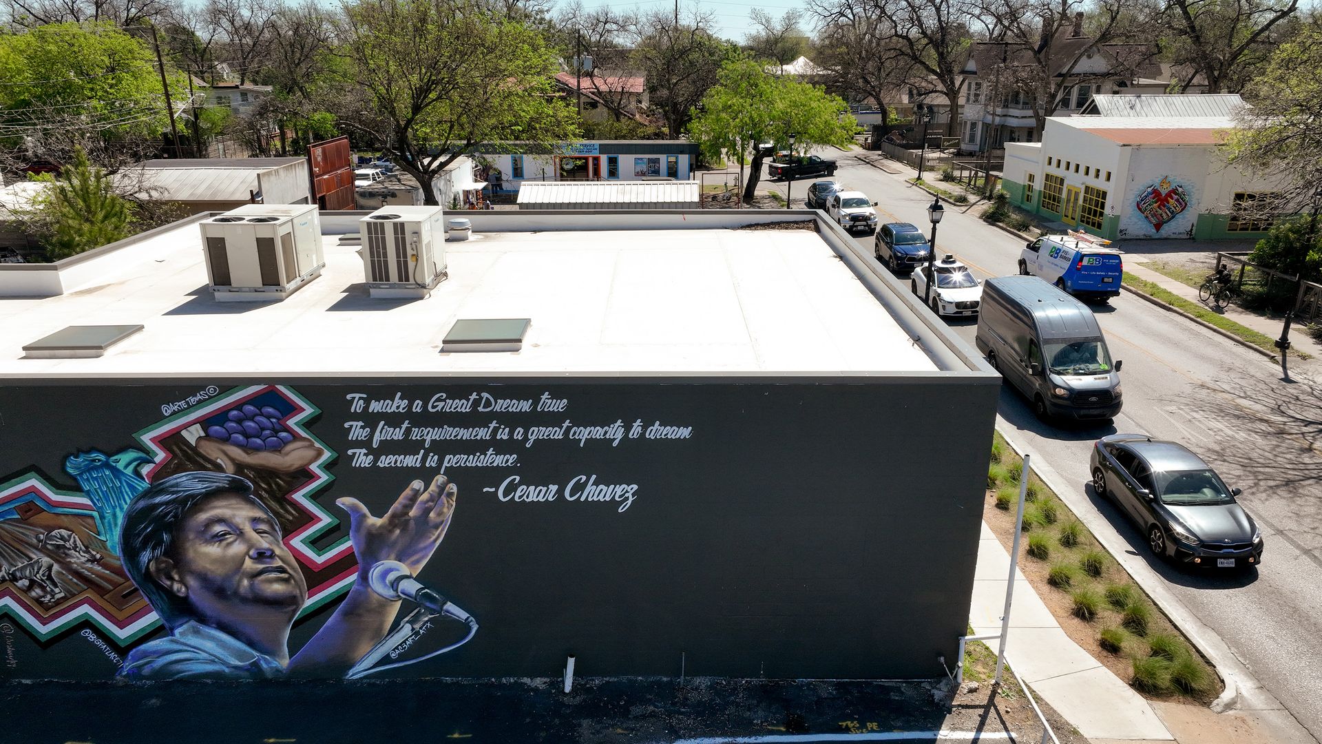 Rooftop mural of Cesar Chavez singing in bold colors with graffiti art on a building wall. A street with cars and trees runs below under a clear blue sky.