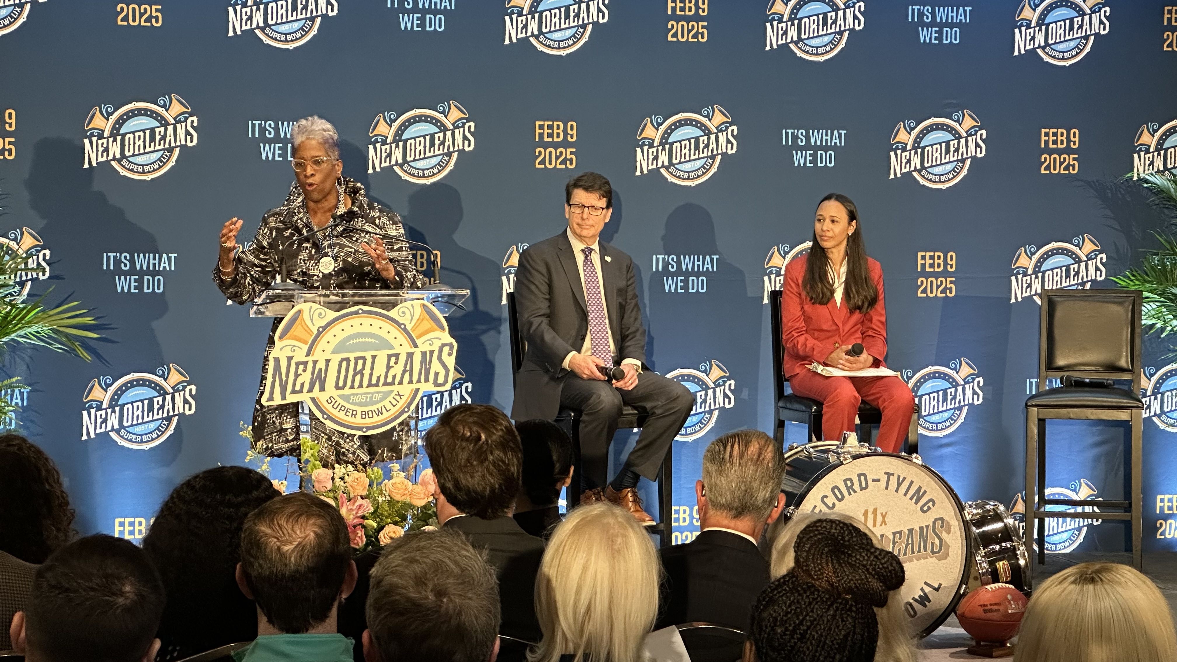 Photo shows three people on state with New Orleans Super Bowl branding behind them.