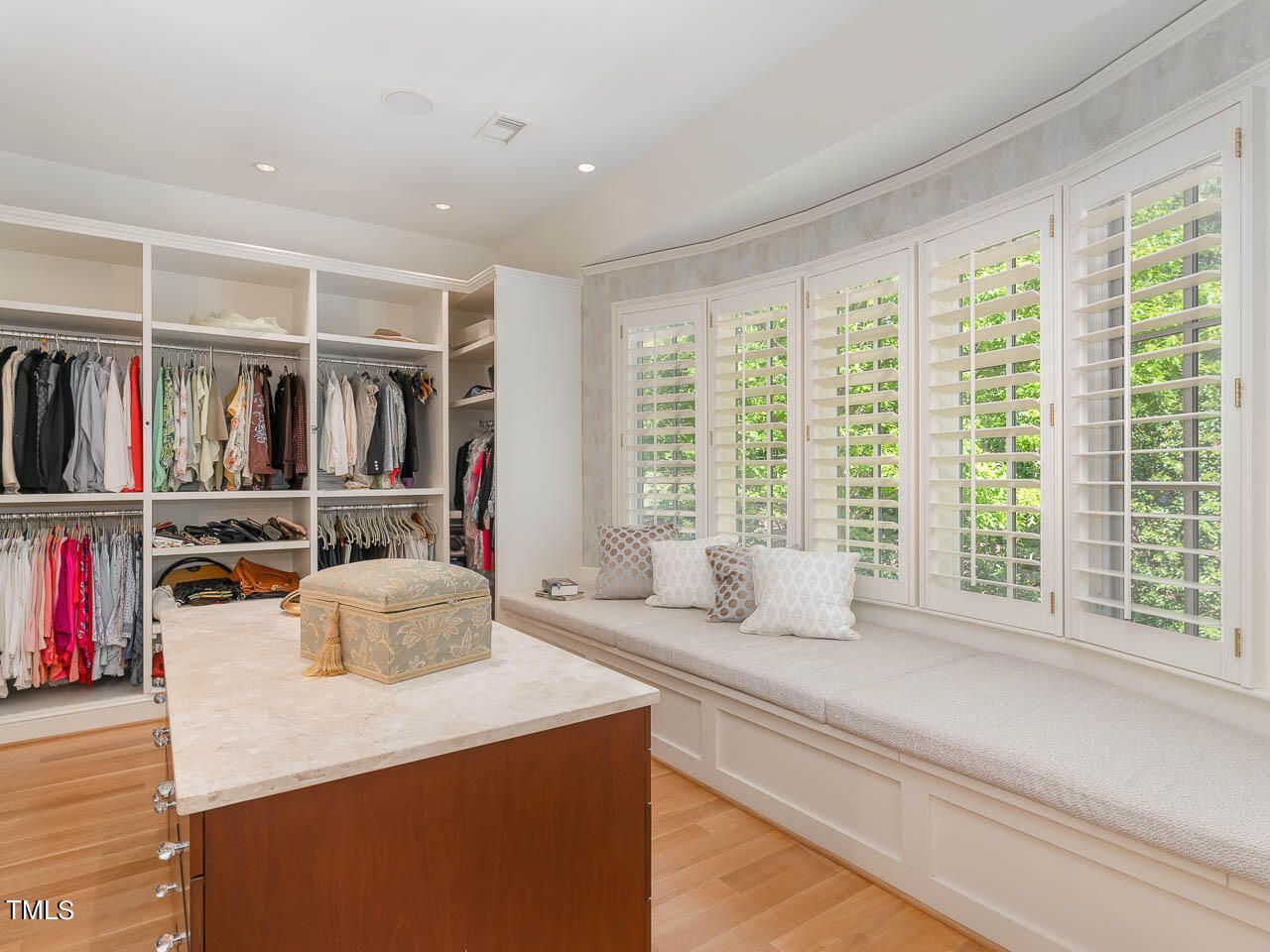 Bright walk-in closet with wooden floor, white built-in shelves holding clothes, a marble-topped island, and a window seat with cushions by white shuttered windows showing green foliage outside.