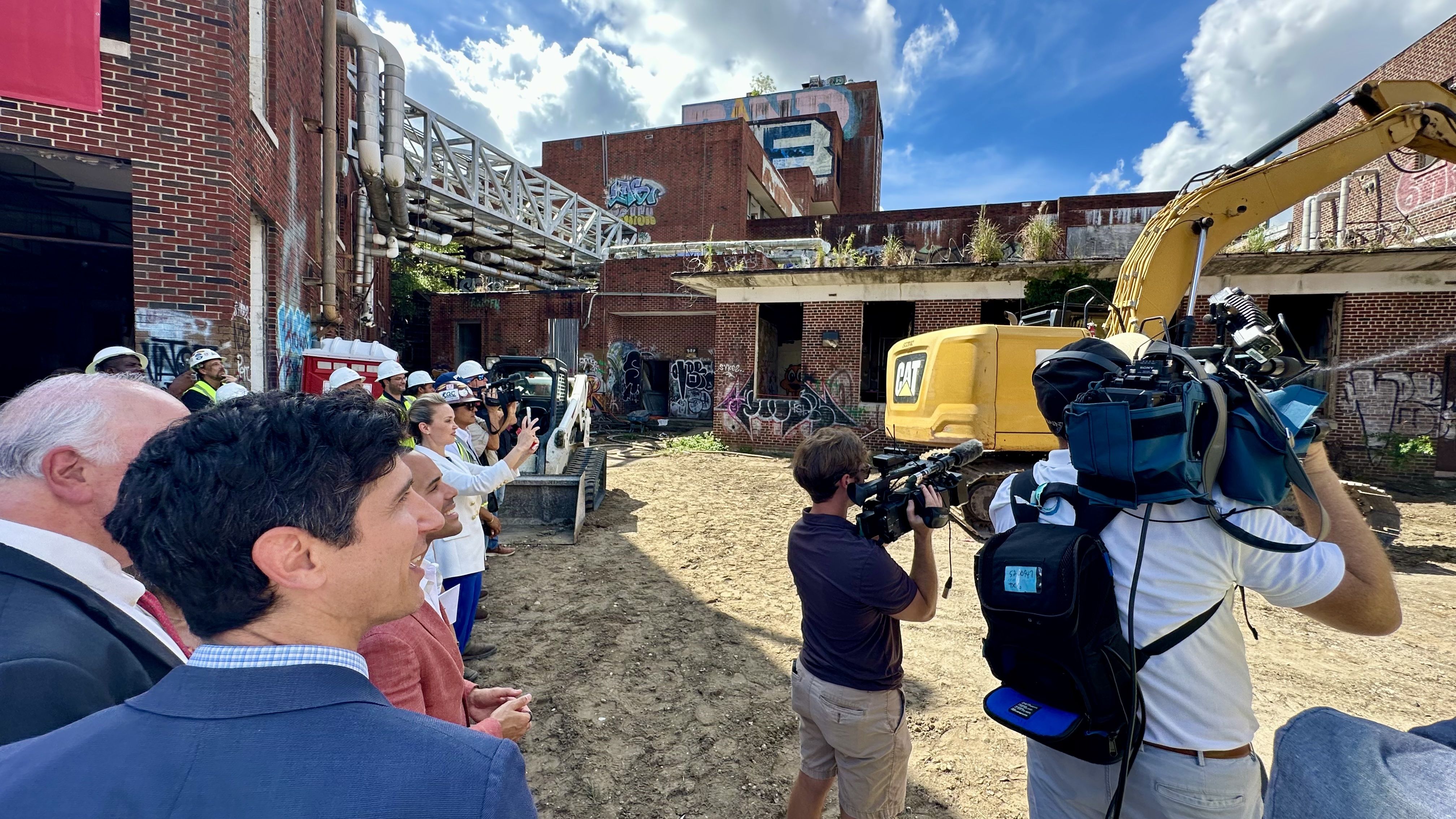 A group of reporters and construction workers wearing hard hats stand in front of a graffiti-covered urban demolition site. A yellow CAT excavator sits among brick buildings and blue sky.