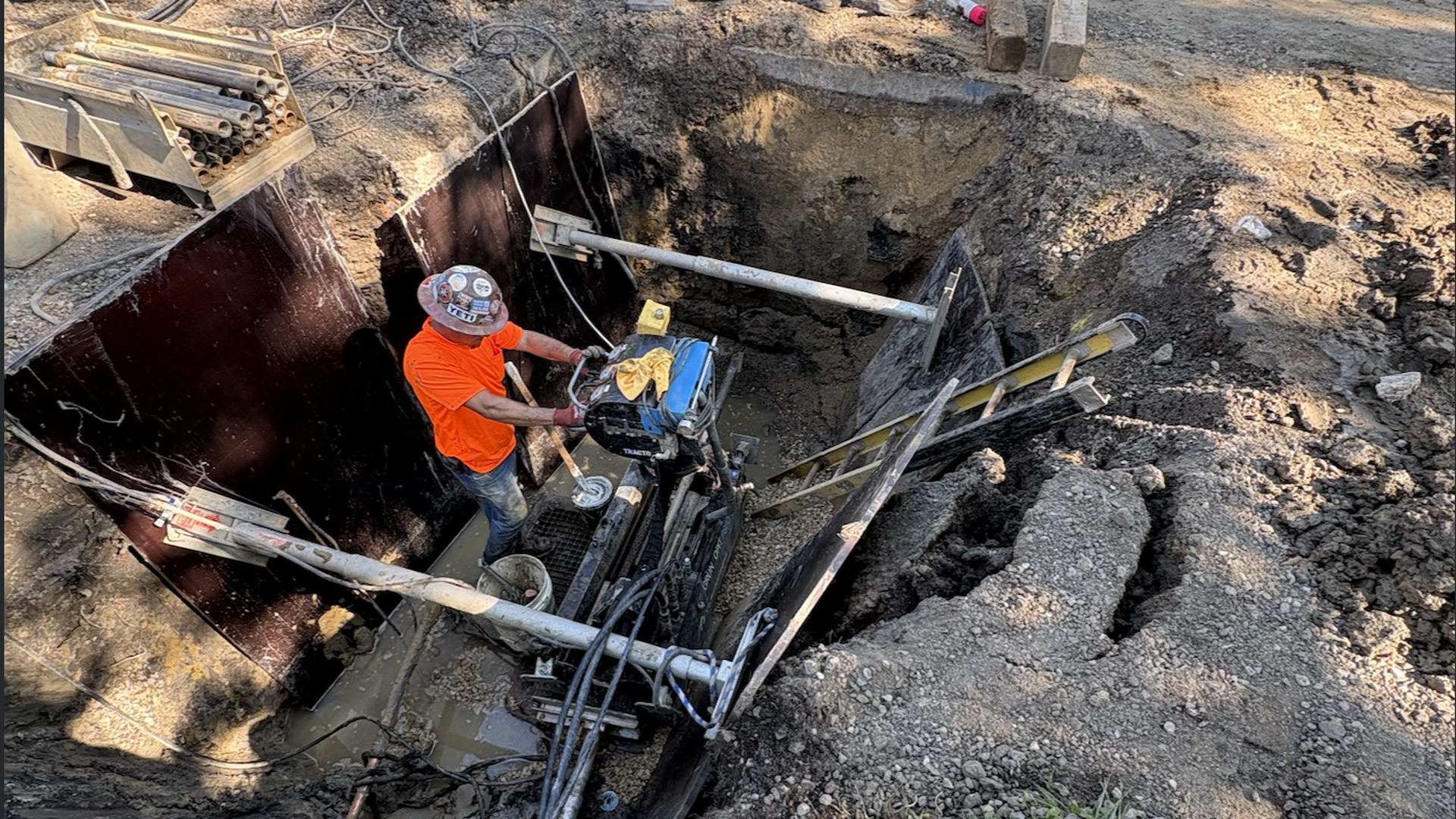 A construction worker in an orange shirt and hard hat operates a compactor at the bottom of a muddy trench, surrounded by steel plates, pipes, ladders, and tangled cables.