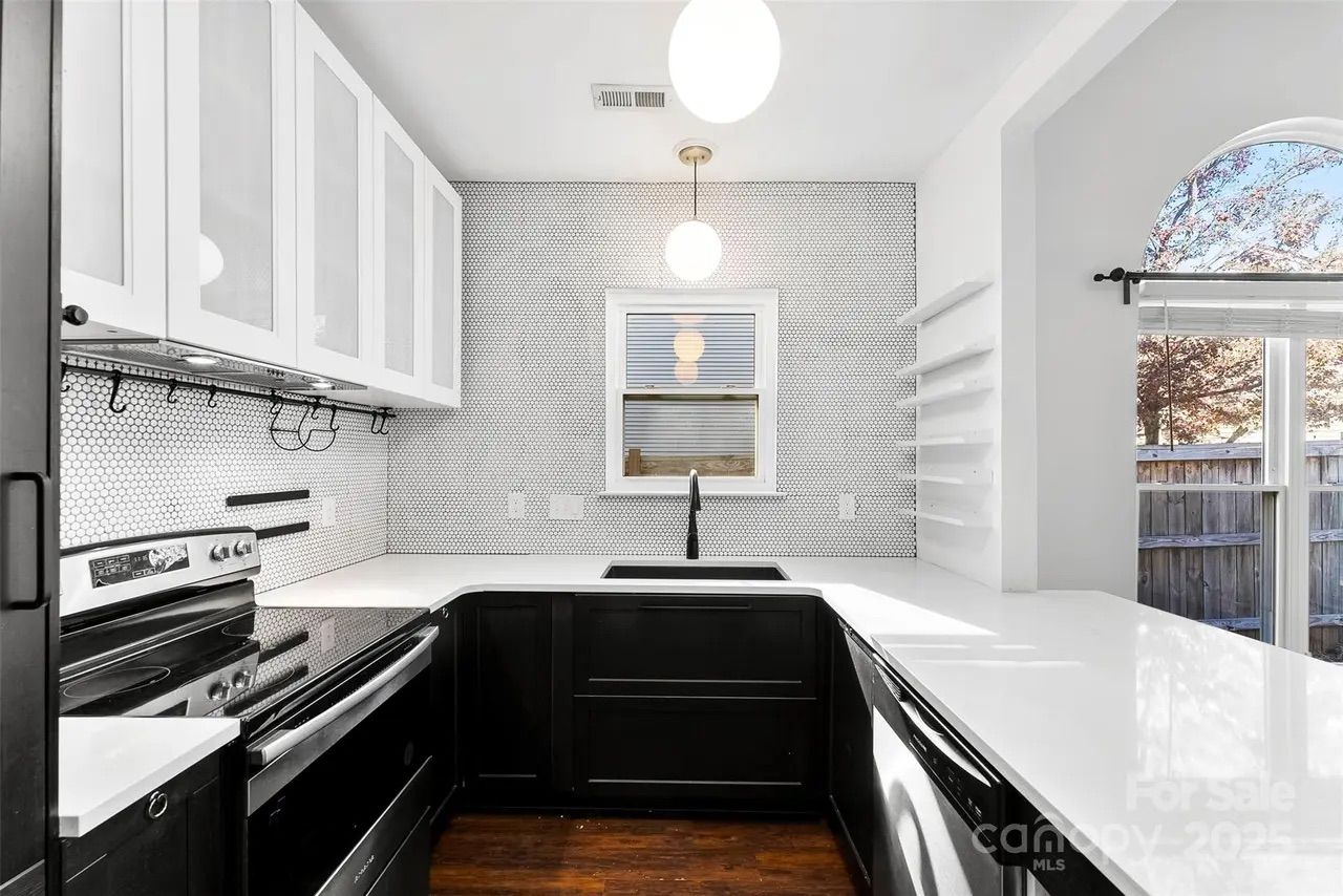 Modern kitchen with black lower cabinets, white upper cabinets, white countertops, small hexagonal white tile backsplash, black faucet, stainless steel stove and dishwasher, and a window above sink.