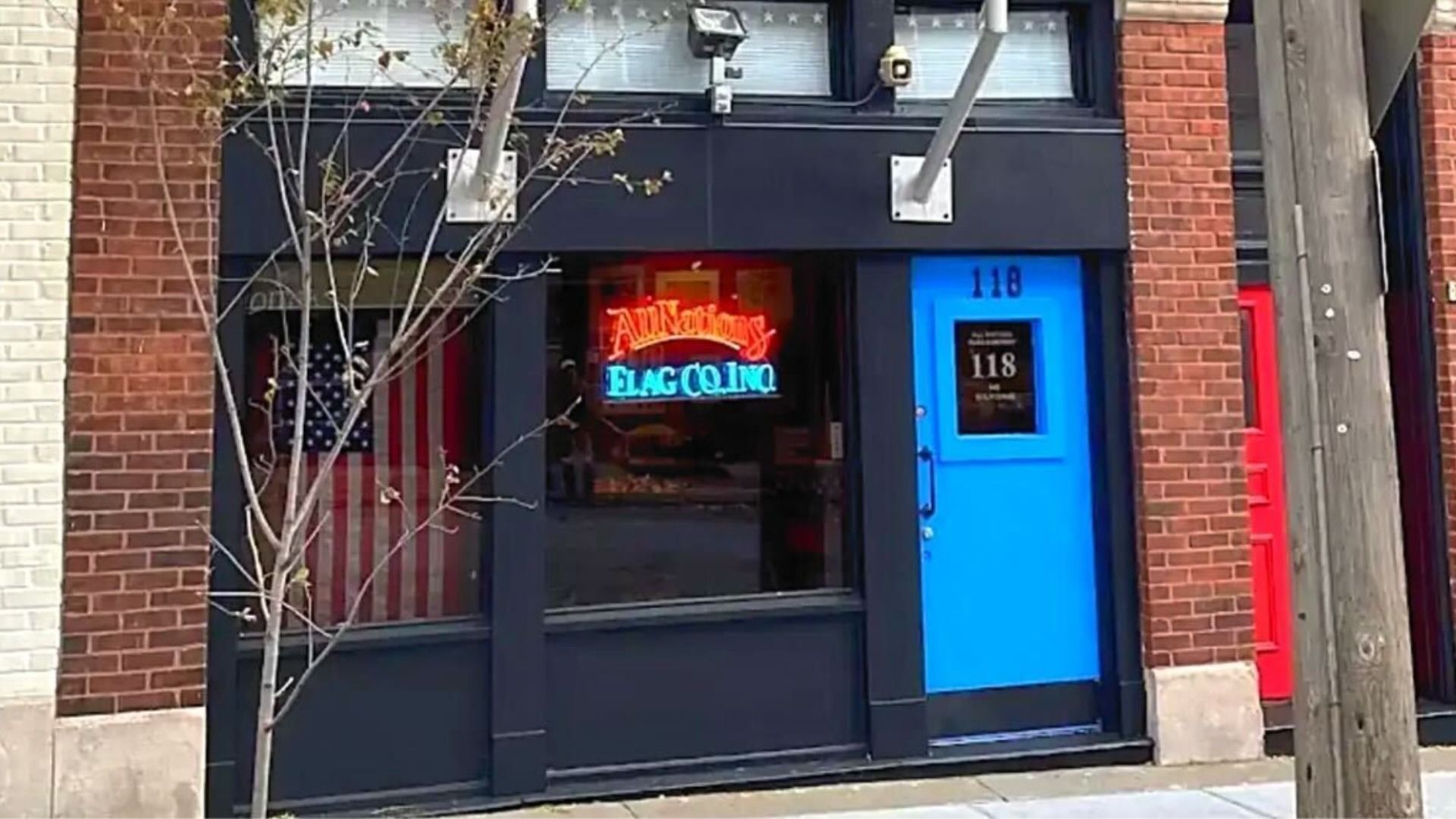 Front view of a brick building with a blue door numbered 118 and a neon sign in the window reading "All Nations Flag Co. Inc." An American flag is visible inside. A small tree stands outside.
