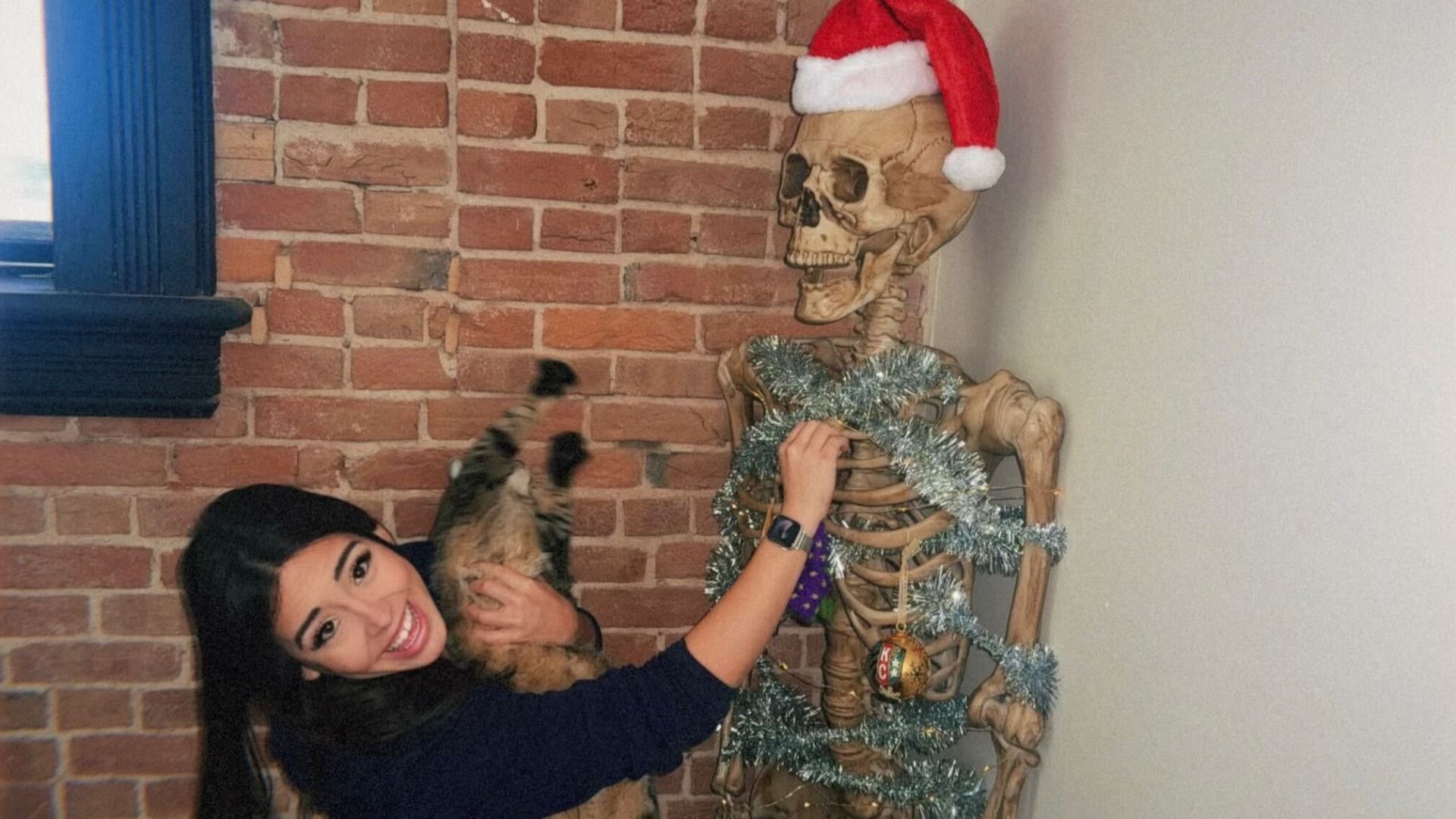 Smiling woman holding a cat, decorating a skeleton with silver tinsel and Christmas ornaments. Skeleton wears a red Santa hat, standing against a brick wall near a window.