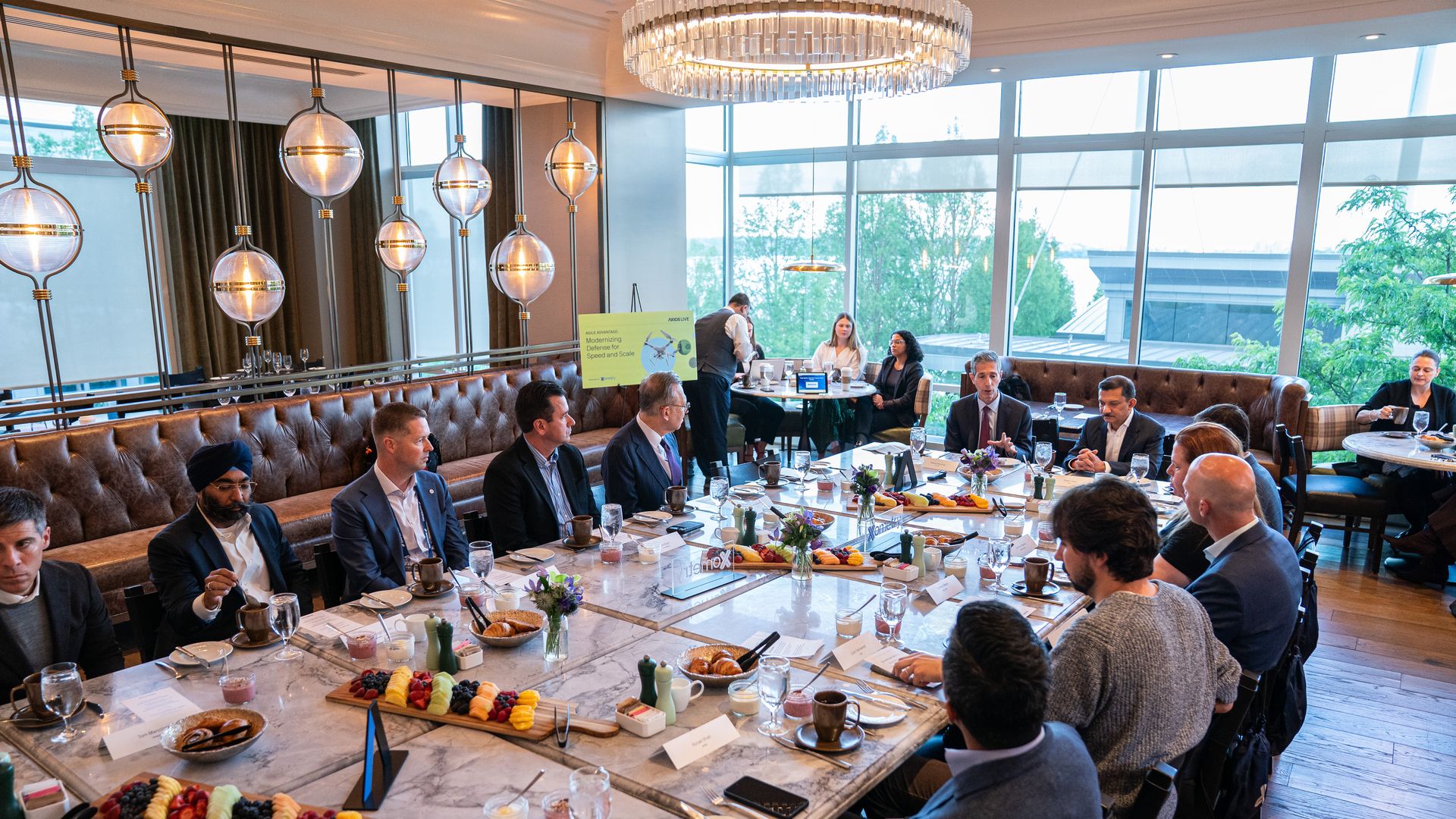 Business meeting in a bright restaurant: professionals in suits sit around a long marble table with plates of fruit, pastries, coffee, and glassware. Glass chandeliers and windows overlook greenery.