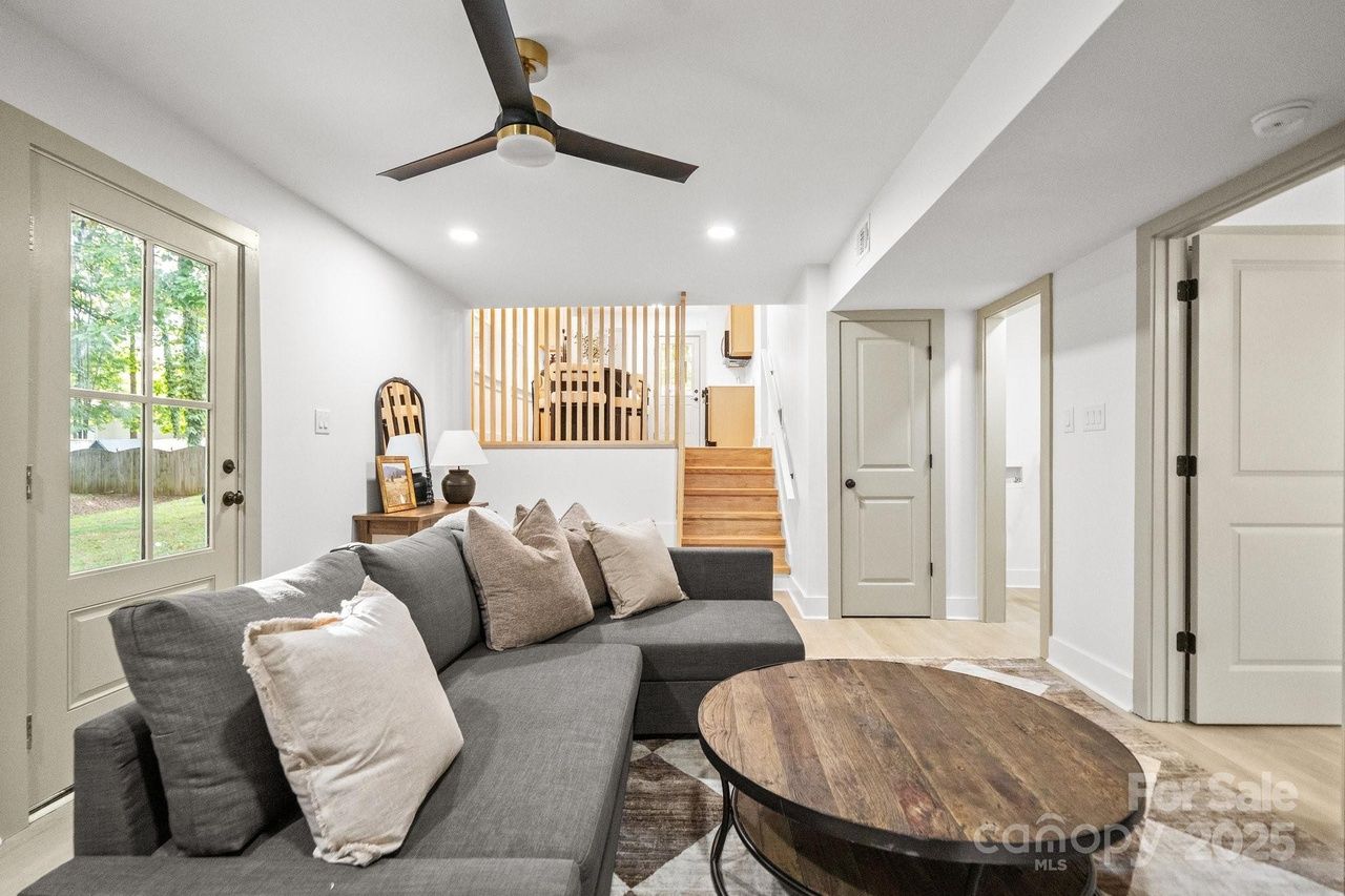 Bright living room with gray sectional sofa, beige cushions, wooden round coffee table, ceiling fan, and stairs leading to a dining area separated by vertical wooden slats.