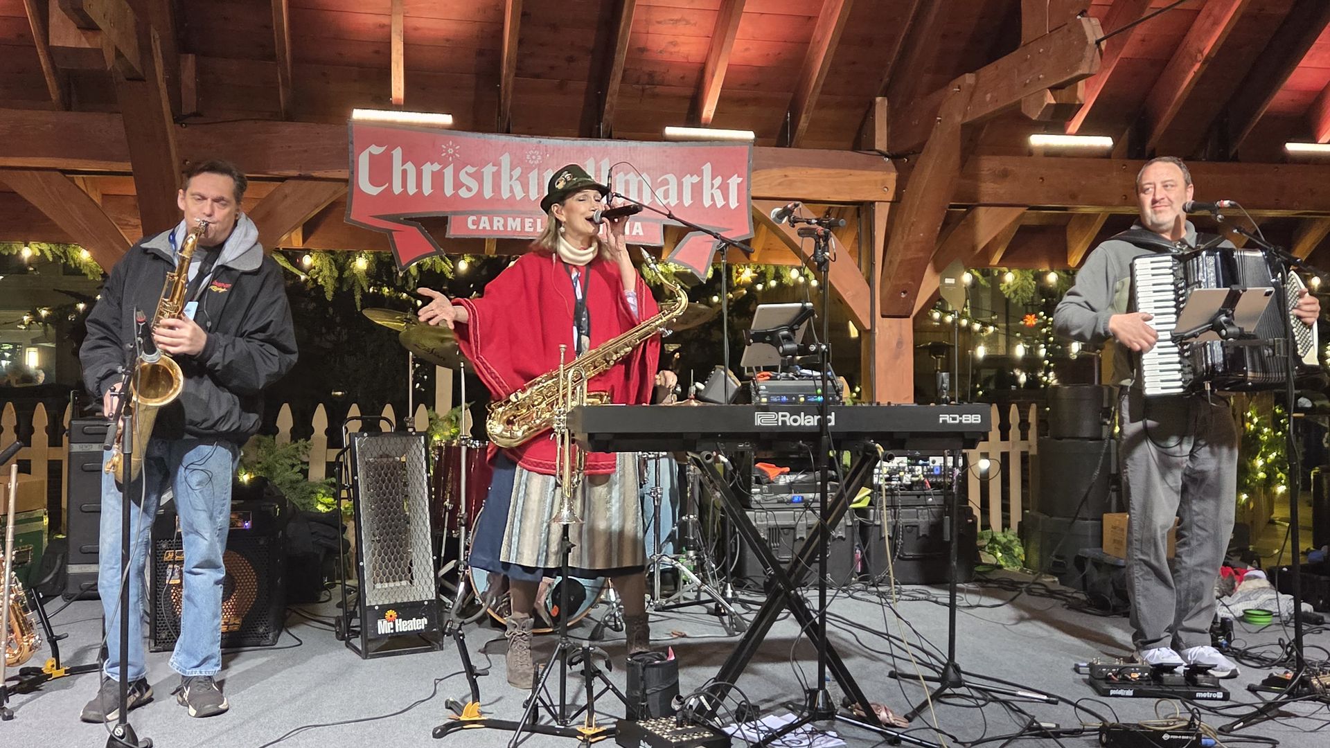 Three musicians performing on a wooden stage under a roof at Christkindlmarkt Carmel, USA. Man on left plays saxophone, woman in center sings with saxophone, man on right plays accordion.