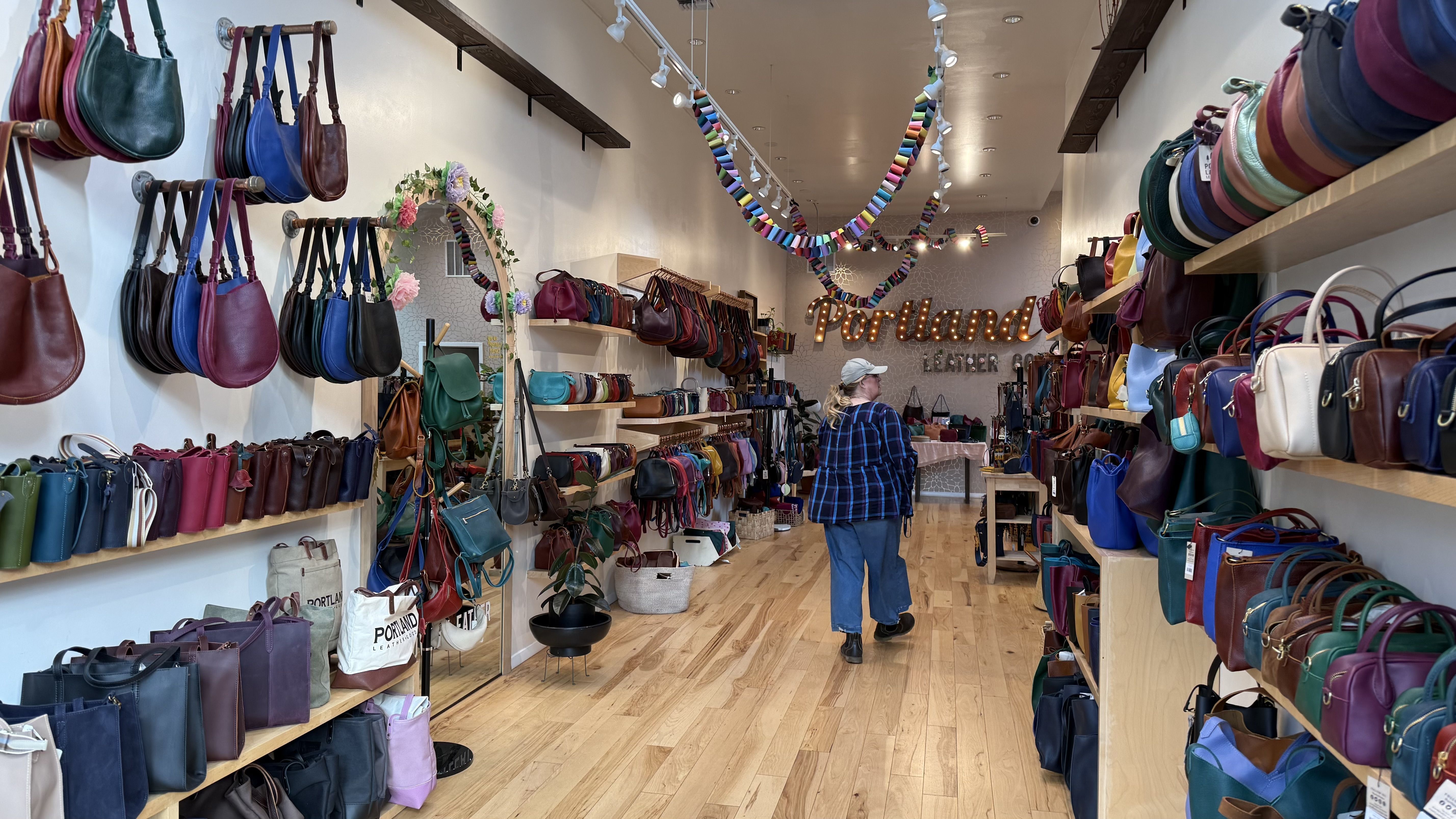 Interior of a leather bag store with wooden shelves filled with colorful handbags. A person in a blue plaid shirt walks inside. A lighted sign reads "Portland Leather Co."