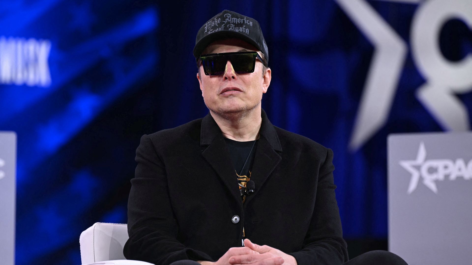 Elon Musk sits on a stage at CPAC wearing sunglasses and a hat.