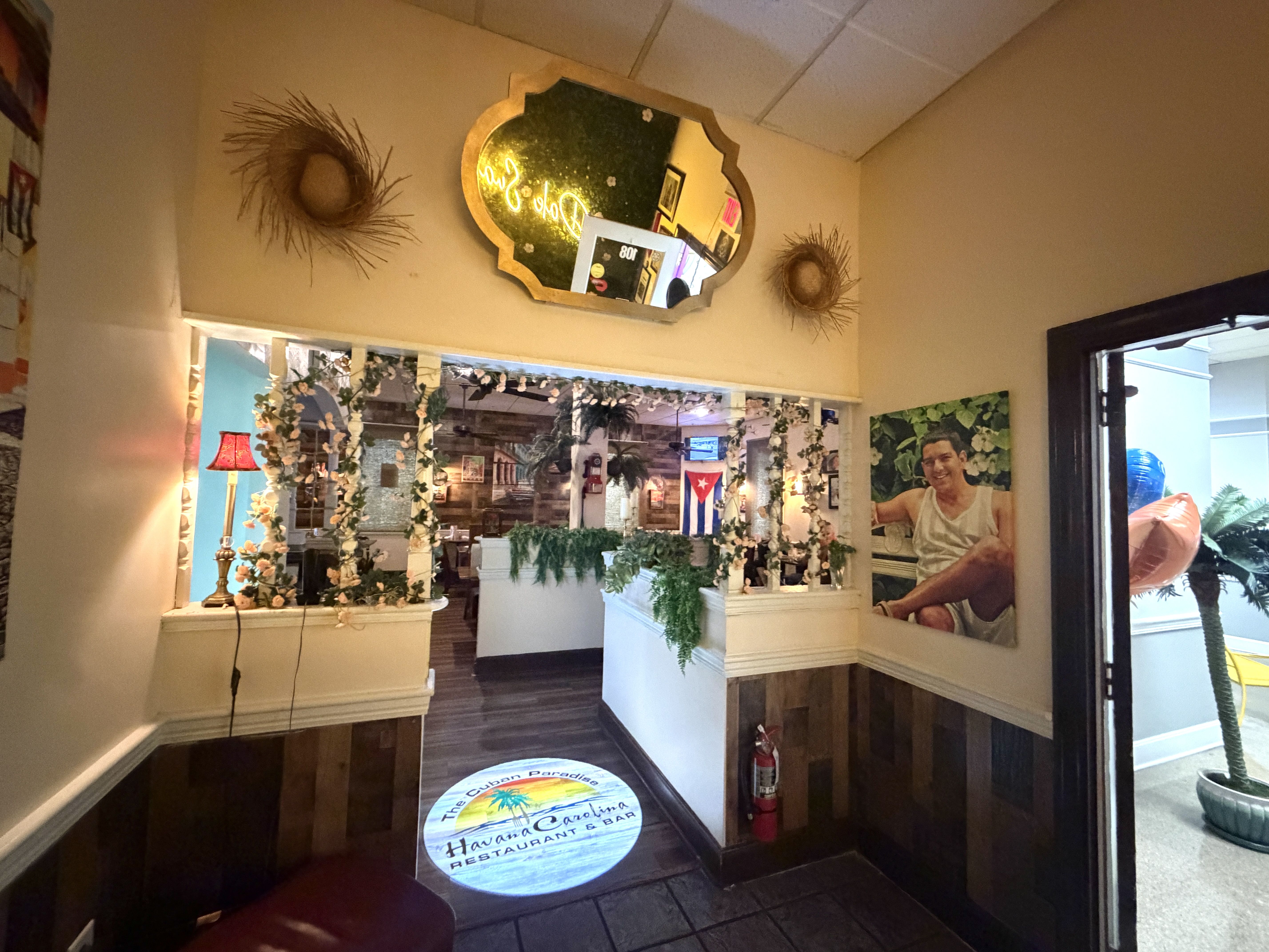 Interior of a Cuban restaurant with beige walls, a gold-framed mirror, two sunburst wicker wall nests, leafy garlands, and circular floor decals reading The Cuban Paradise.