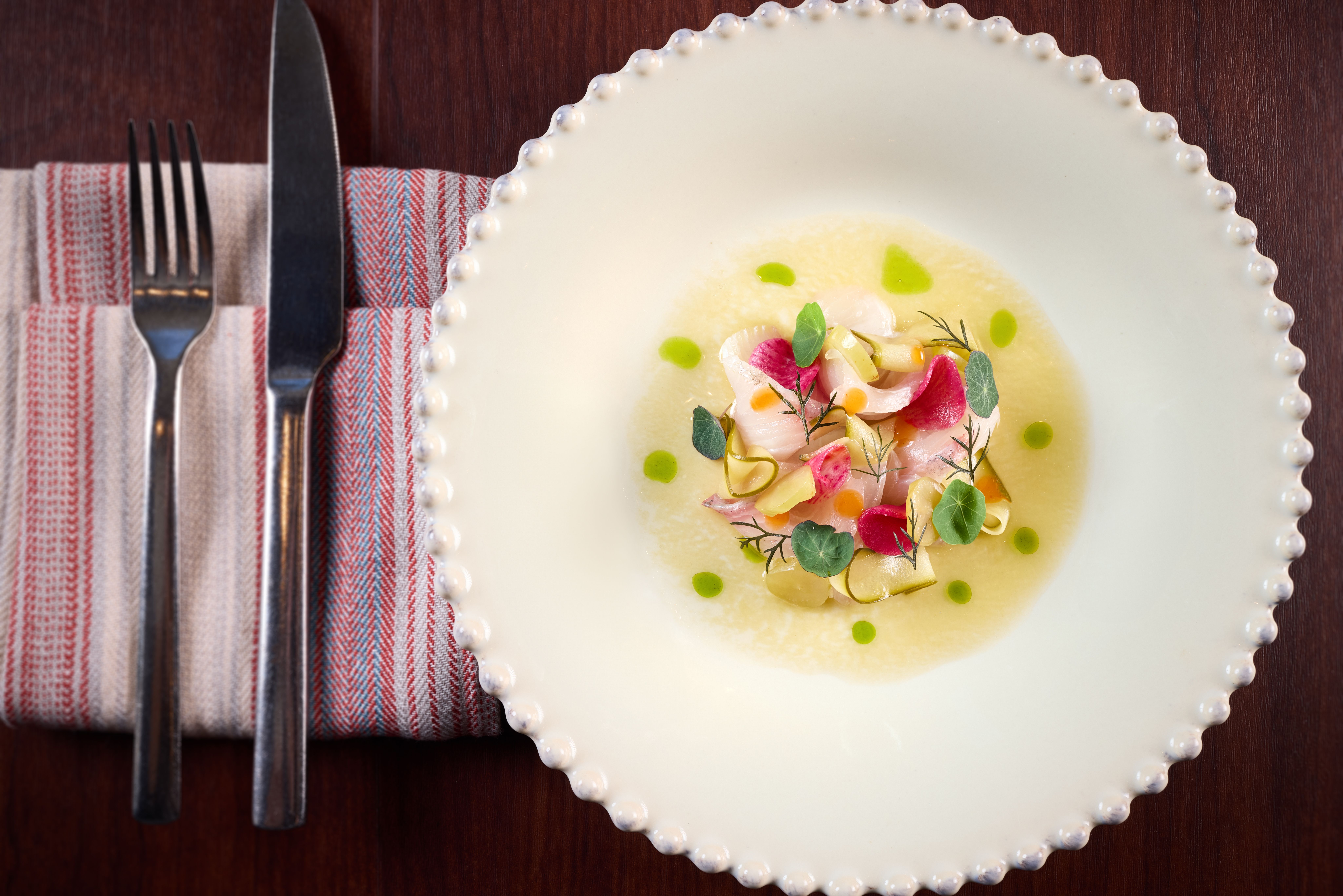 Center-plated fine-dining dish: colorful vegetables and fish on pale yellow sauce, microgreens garnish, white beaded-edge plate on dark wood with striped napkin and silverware.