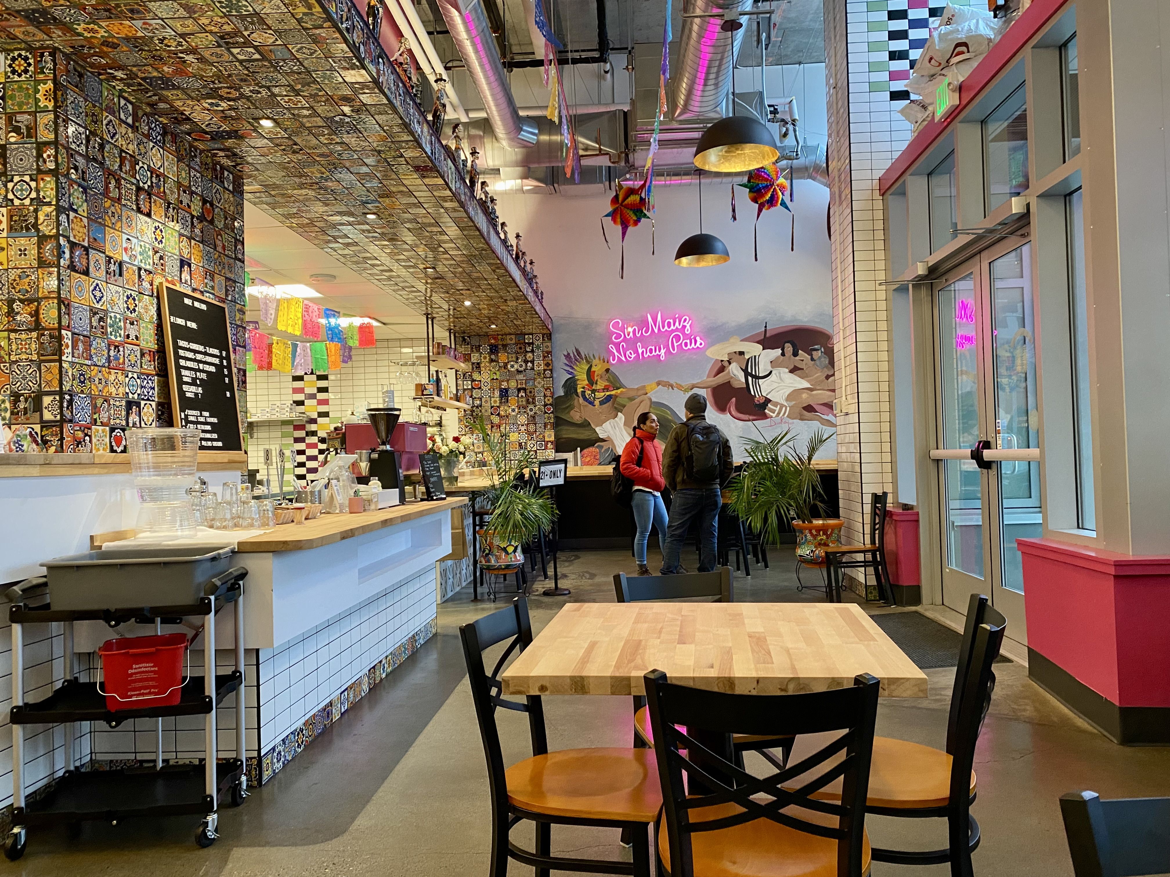 A view of the interior of a restaurant with colorful tiles on the wall, a counter at the left, a fourtop table at center and two people talking in the background. 