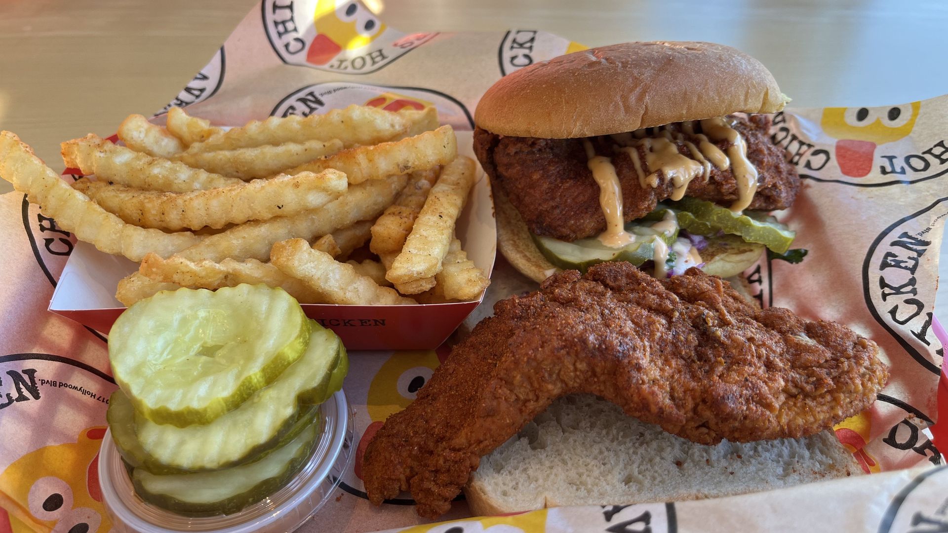 A hot chicken sandwich next to a pile of fries and a cup of pickles