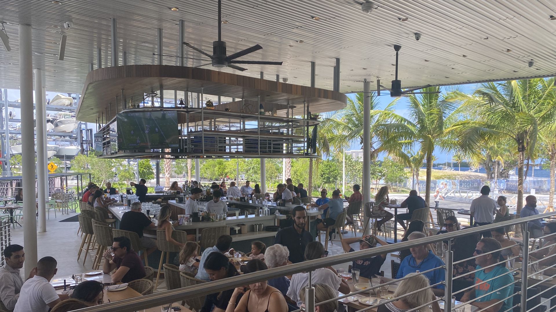 Diners gather at tables around a large bar, with palm trees in the background on a sunny afternoon. 