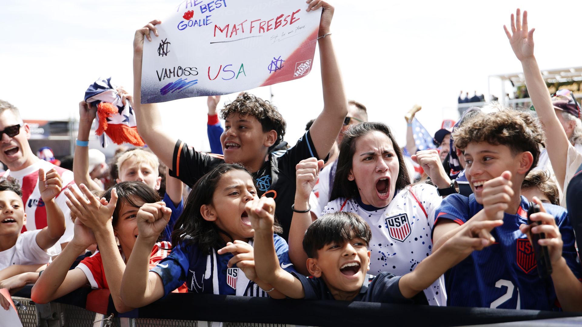 Soccer fans wearing blue, white and red Team USA jerseys cheer and raise their hands in a crowd.