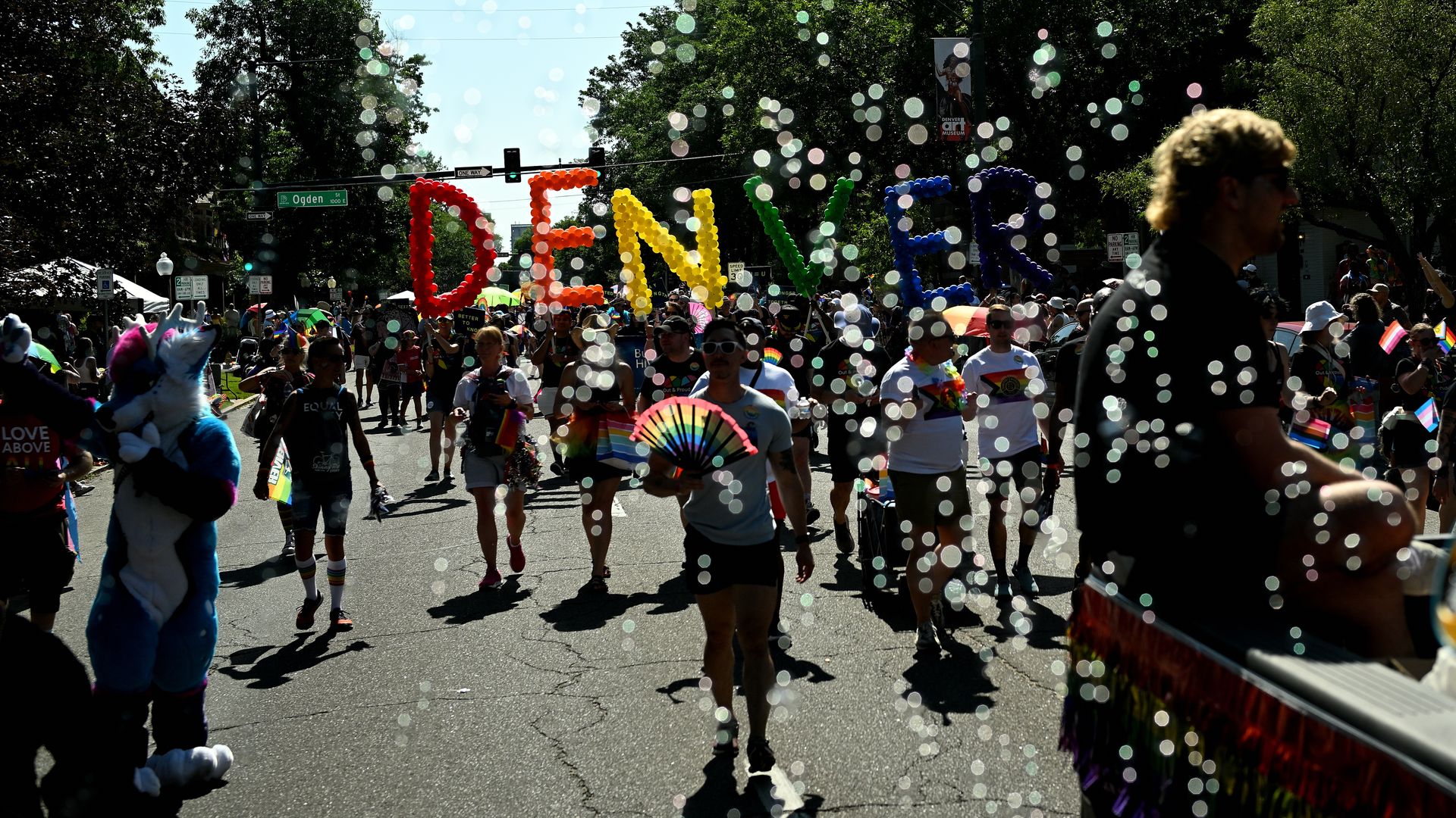 People in colorful attire participate in a Denver pride parade holding large balloon letters spelling "DENVER" in red, orange, yellow, green, blue, and purple, with rainbow flags.