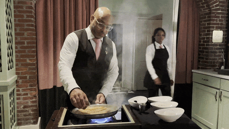 Image shows a man making a flambeed bananas Foster on a tableside cart. There's a big flame.