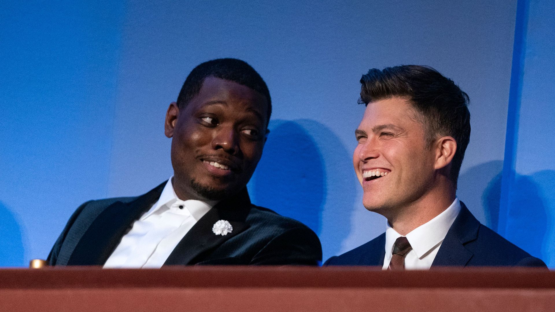 Michael Che, left, looks at Colin Jost, right, who is smiling. 