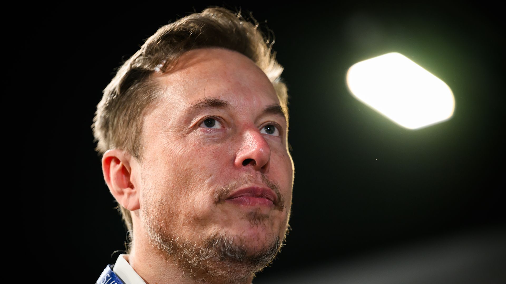 Close-up of a man's face looking upward, showing light stubble, short brown hair, and a blue lanyard with white text around his neck. A bright, blurred light glows in the dark background.