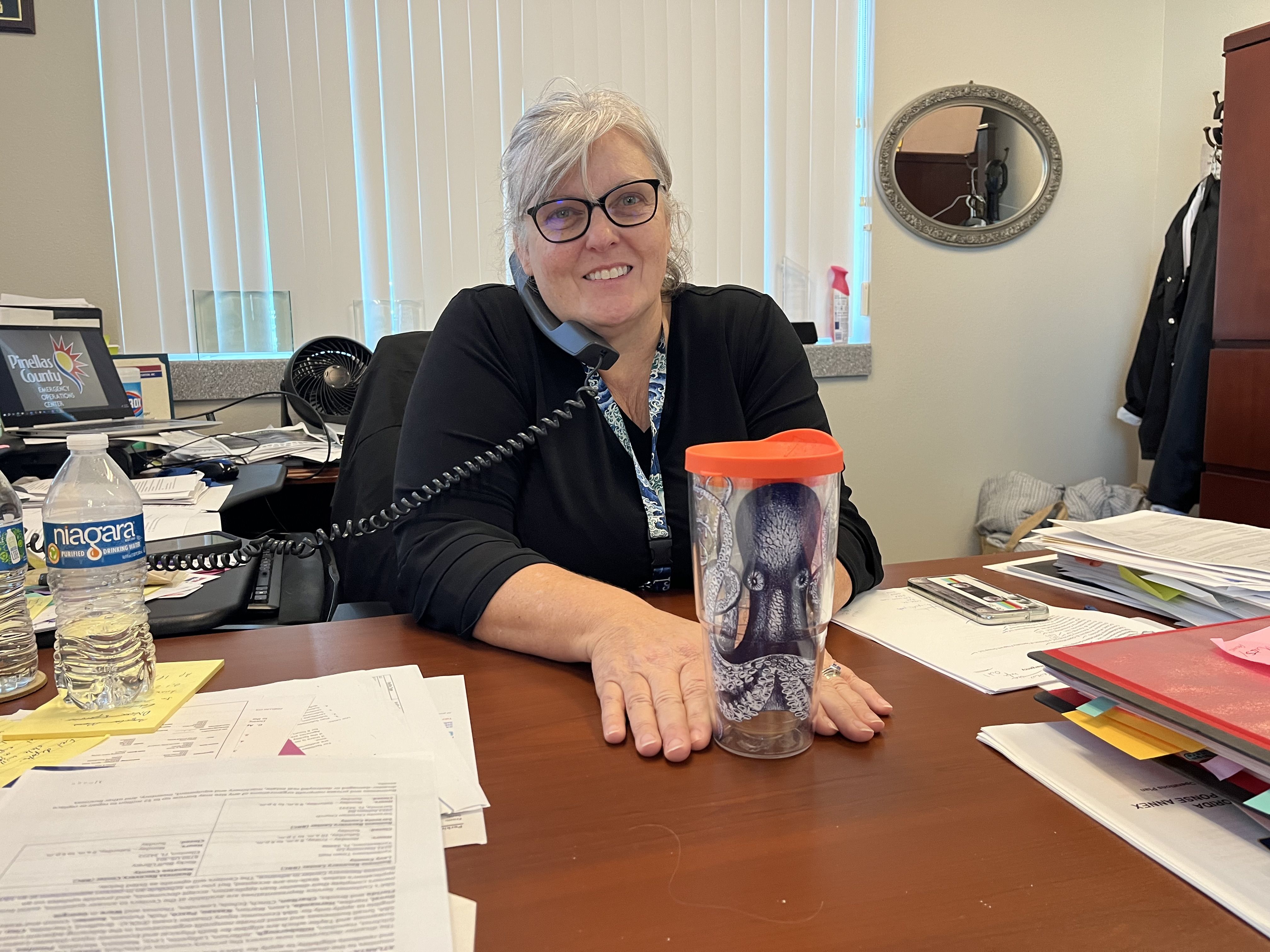 A woman sitting at a desk scattered with papers with a corded phone to he ear shows off a drinking tumbler decorated with a drawing of a blue octopus.