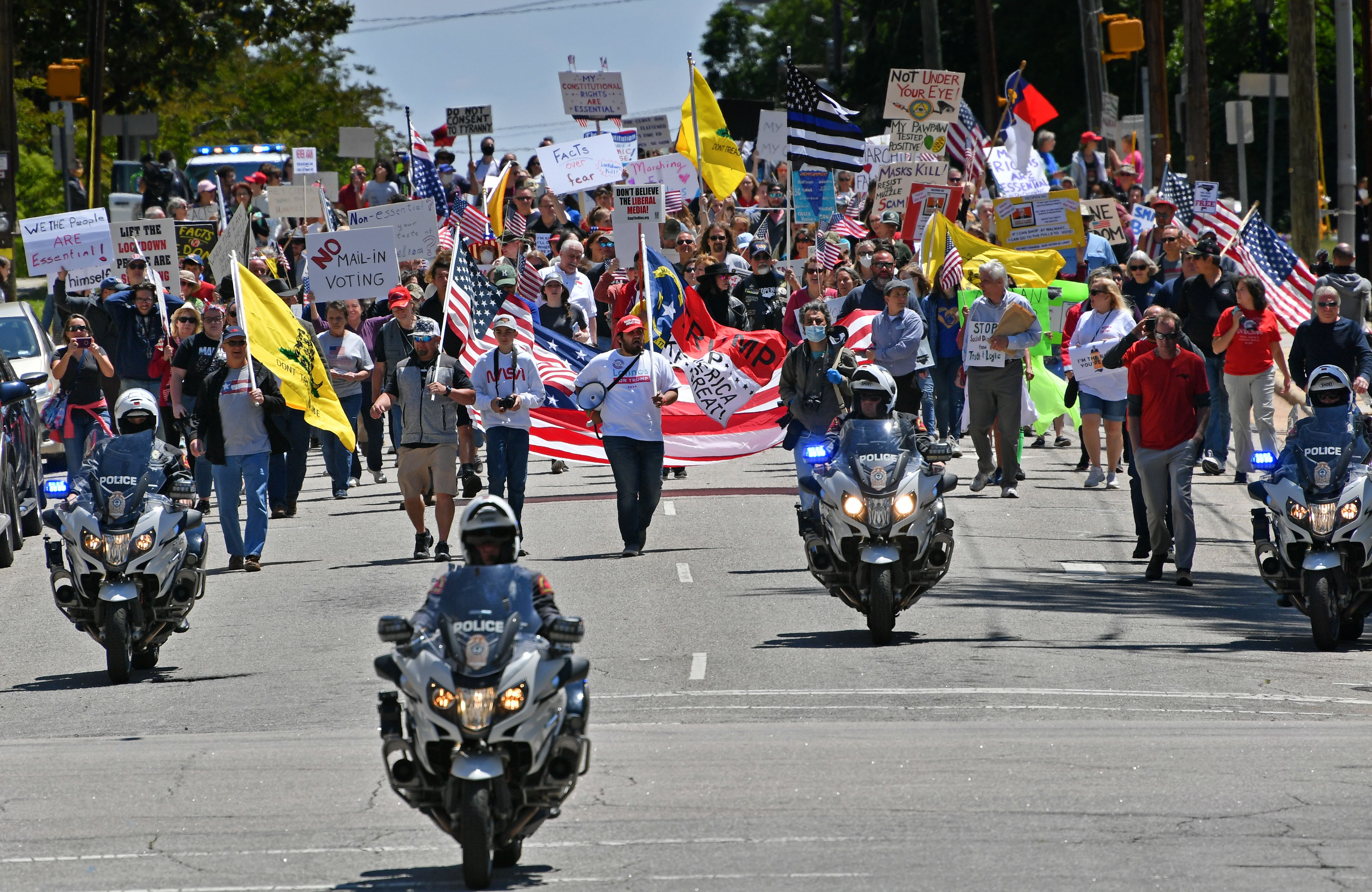 In this image, a large crowd walking down the middle of a street follows a police escort of three motorcycles