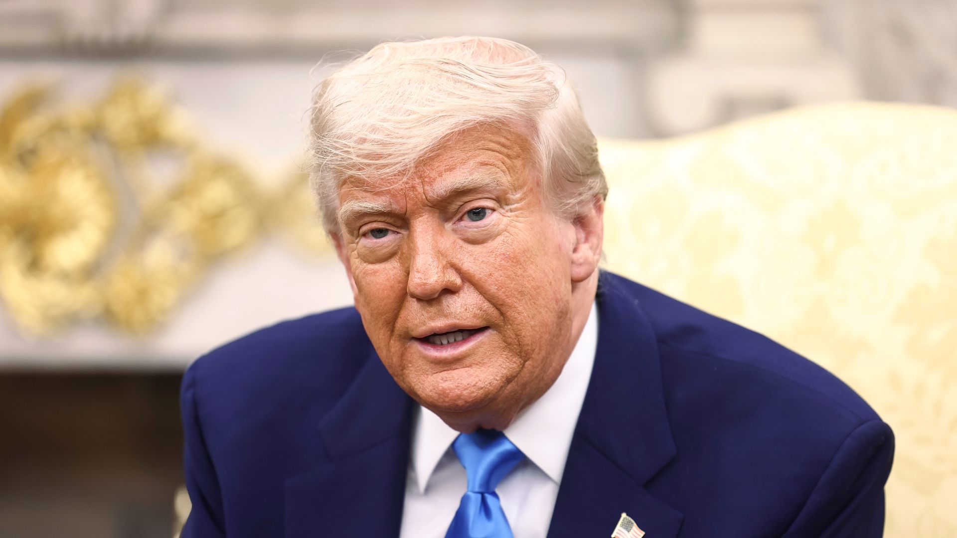 Trump, wearing a blue suit, white shirt and blue tie, sits at a yellow chair 