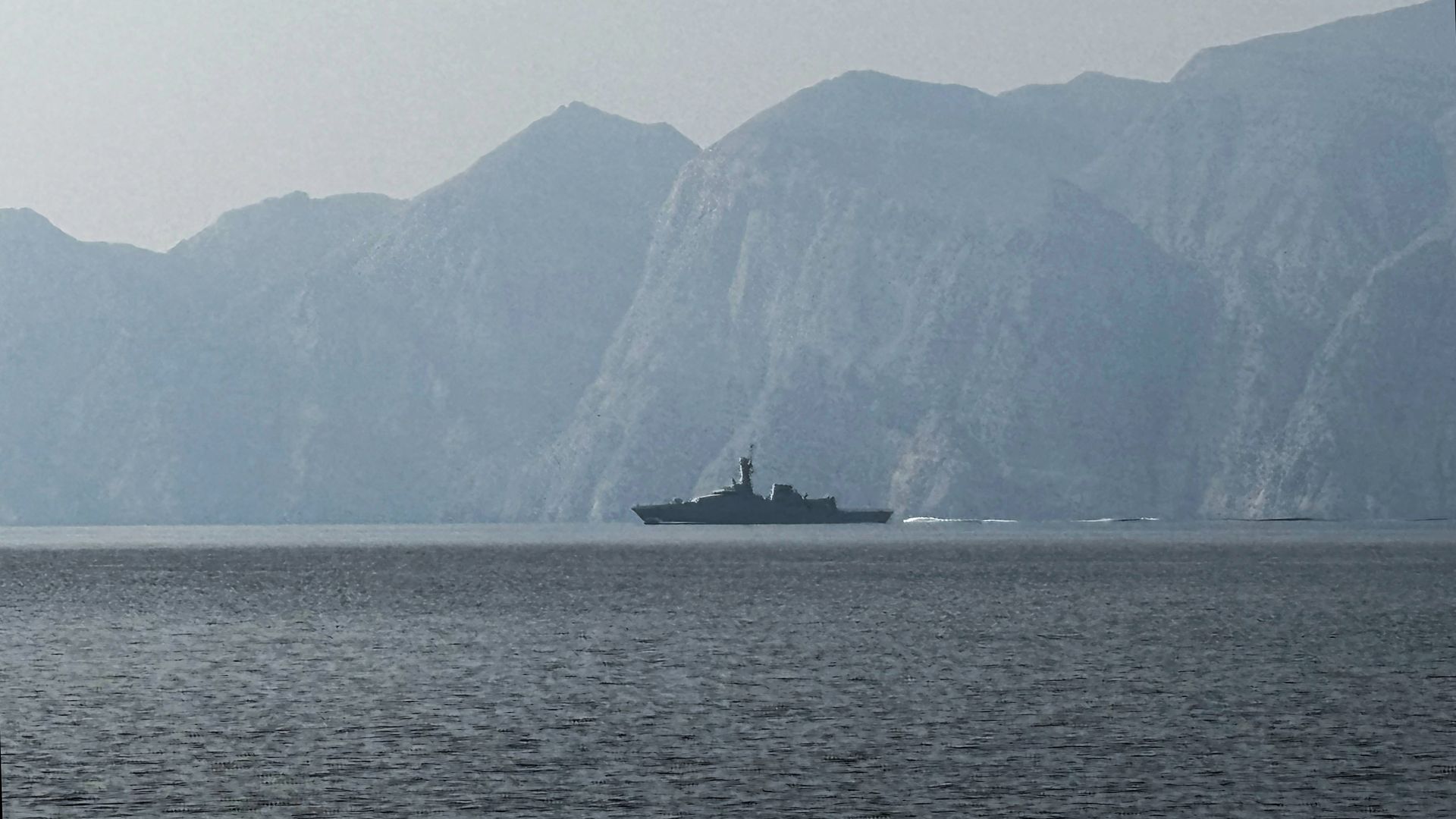 A military ship moves through the Strait of Hormuz with open water and a hazy sky in the background.