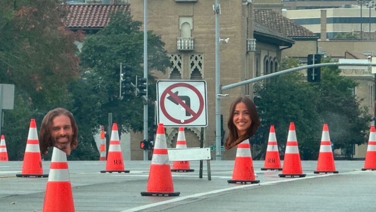 Two orange traffic cones on a road have photos of smiling heads attached, one male and one female, near a no left turn sign and some historic buildings in the background.
