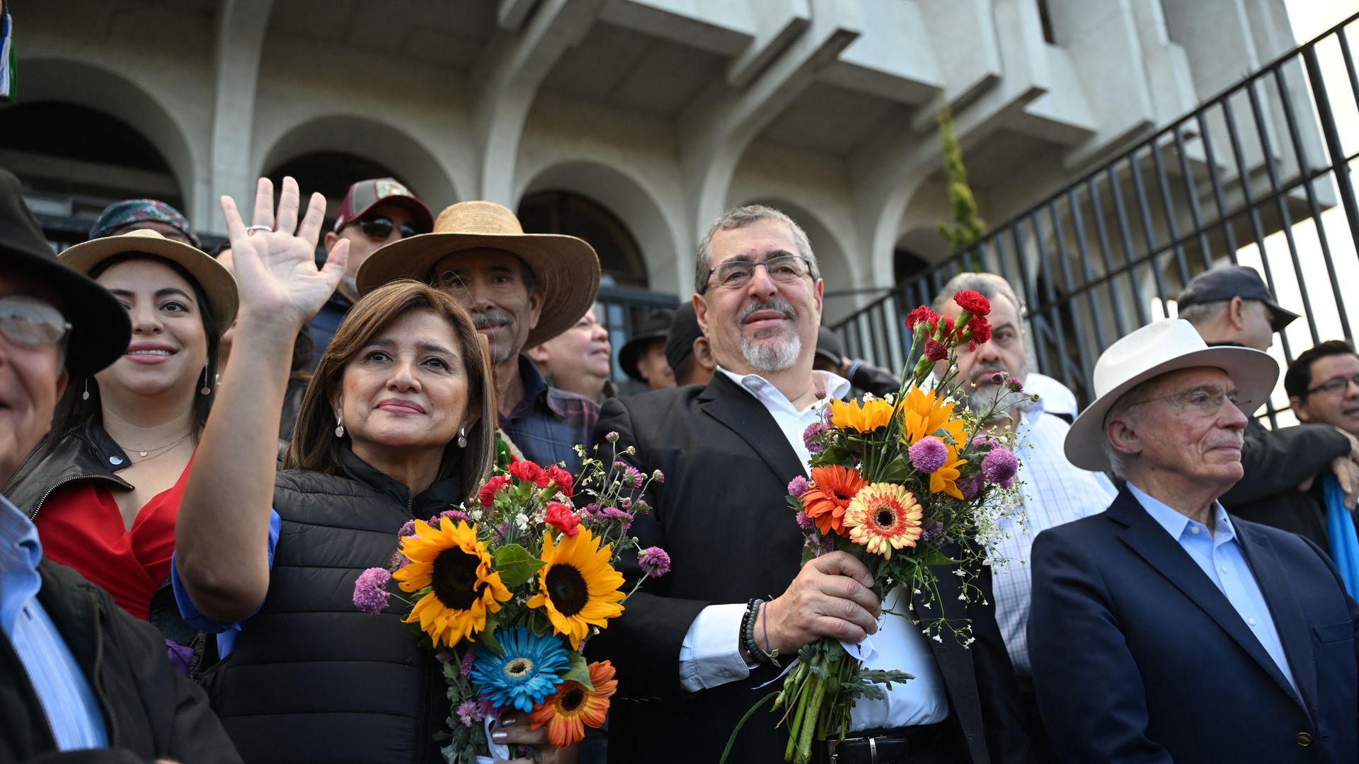 Guatemalan President-elect Bernardo Arévalo and his vice president, Karin Herrera, stand up each holding a bouquet of flowers. they are surrounded by people 