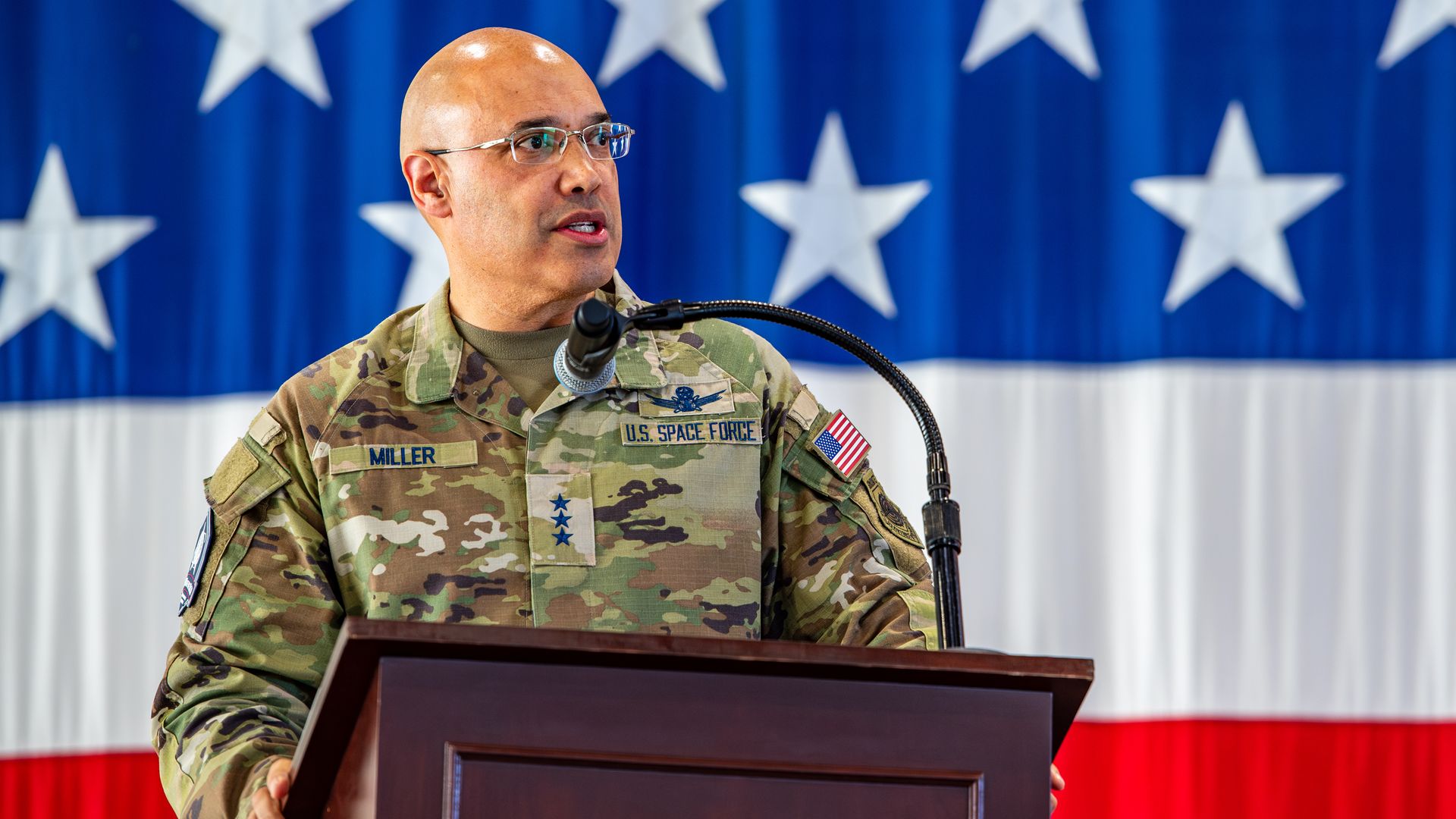 Portrait of a bald, glasses-wearing U.S. Space Force officer at a podium in camouflage.