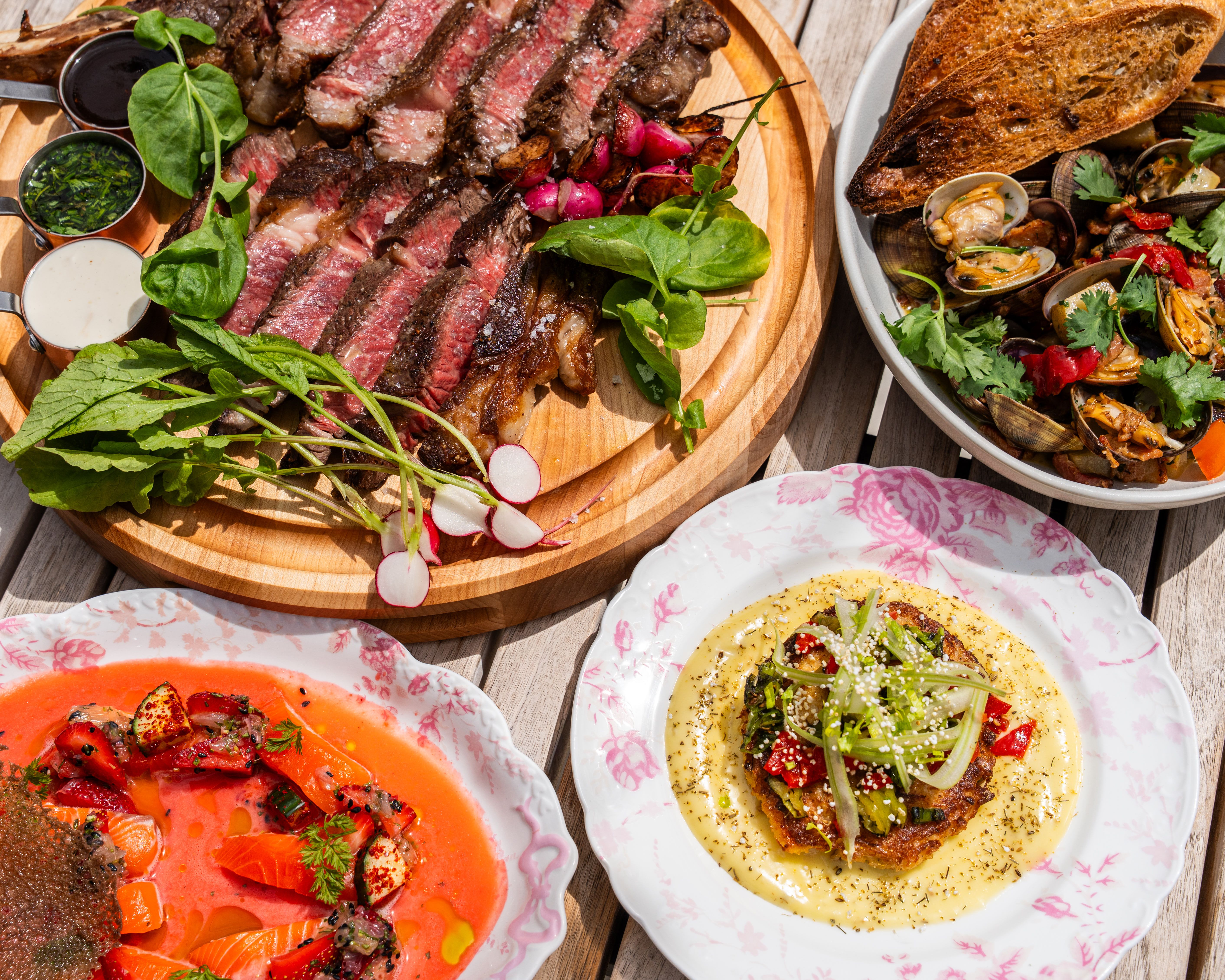 Overhead view of a rustic feast: grilled steak slices with greens on a wooden board, radishes and sauces; beside a pan of clams and mussels with bread, plus salmon and pasta plates.