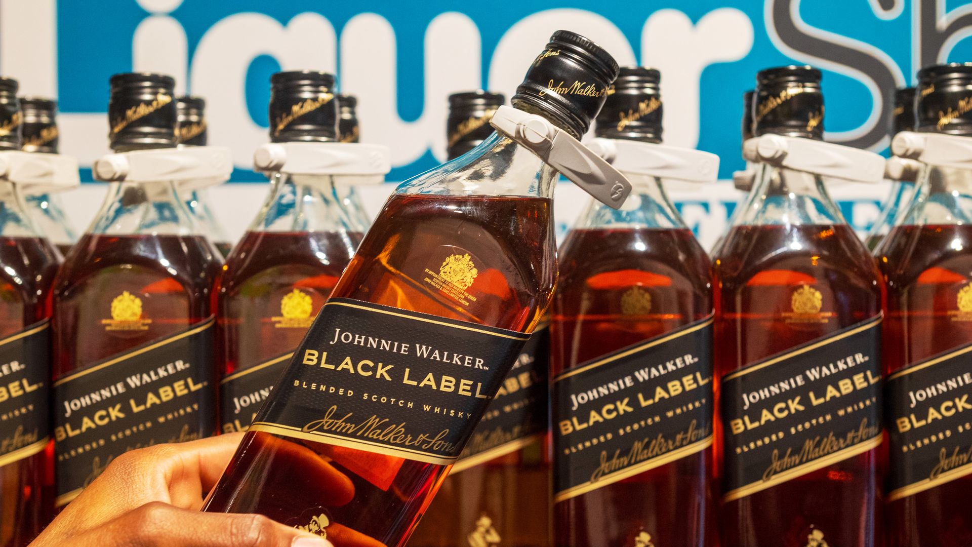 Hand holding a bottle of Johnnie Walker Black Label blended Scotch whisky in front of several similar bottles on display with a partially visible blue and white sign in the background.