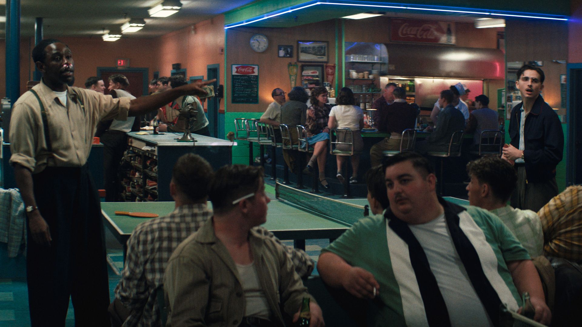 A 1950s scene from "Marty Supreme" with men and women socializing around a ping pong table with a bar in the background. Tyler, The Creator plays a man with a bloody nose pointing at Timothée Chalamet across the table.