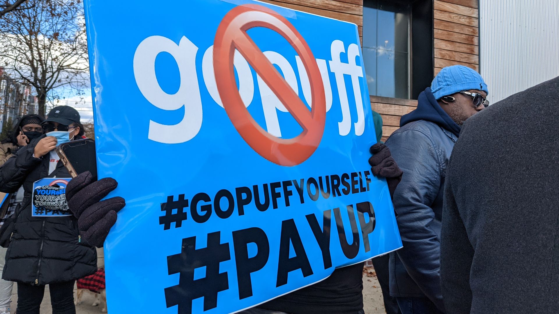 A driver for the delivery company Gopuff strikes outside the company's headquarters holding a sign that reads #gopuffyourself and #payup.