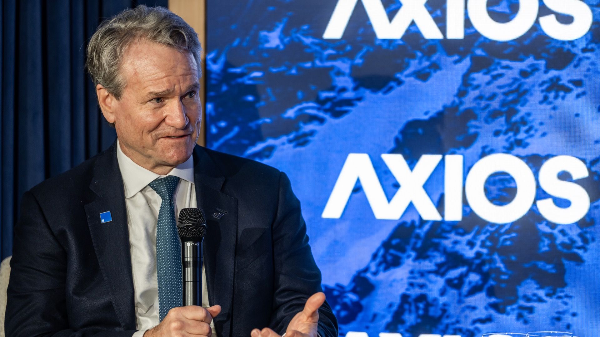 Bank of America CEO in a dark suit and blue tie holding a microphone, speaking in front of a blue backdrop with the word "AXIOS" in white letters.