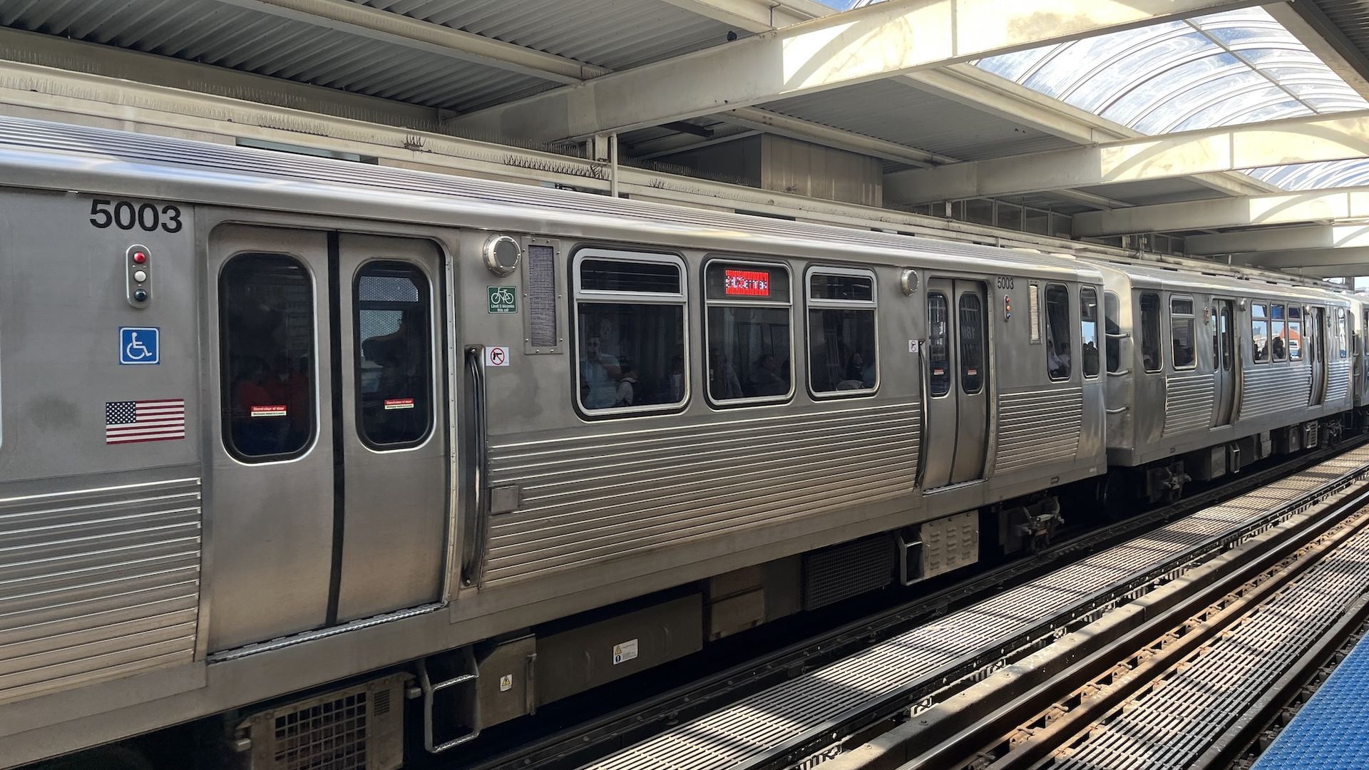 Silver CTA subway train at an elevated station beneath a curved glass canopy. Car 5003 with wheelchair sign and US flag sticker; passengers visible through windows, tracks nearby.