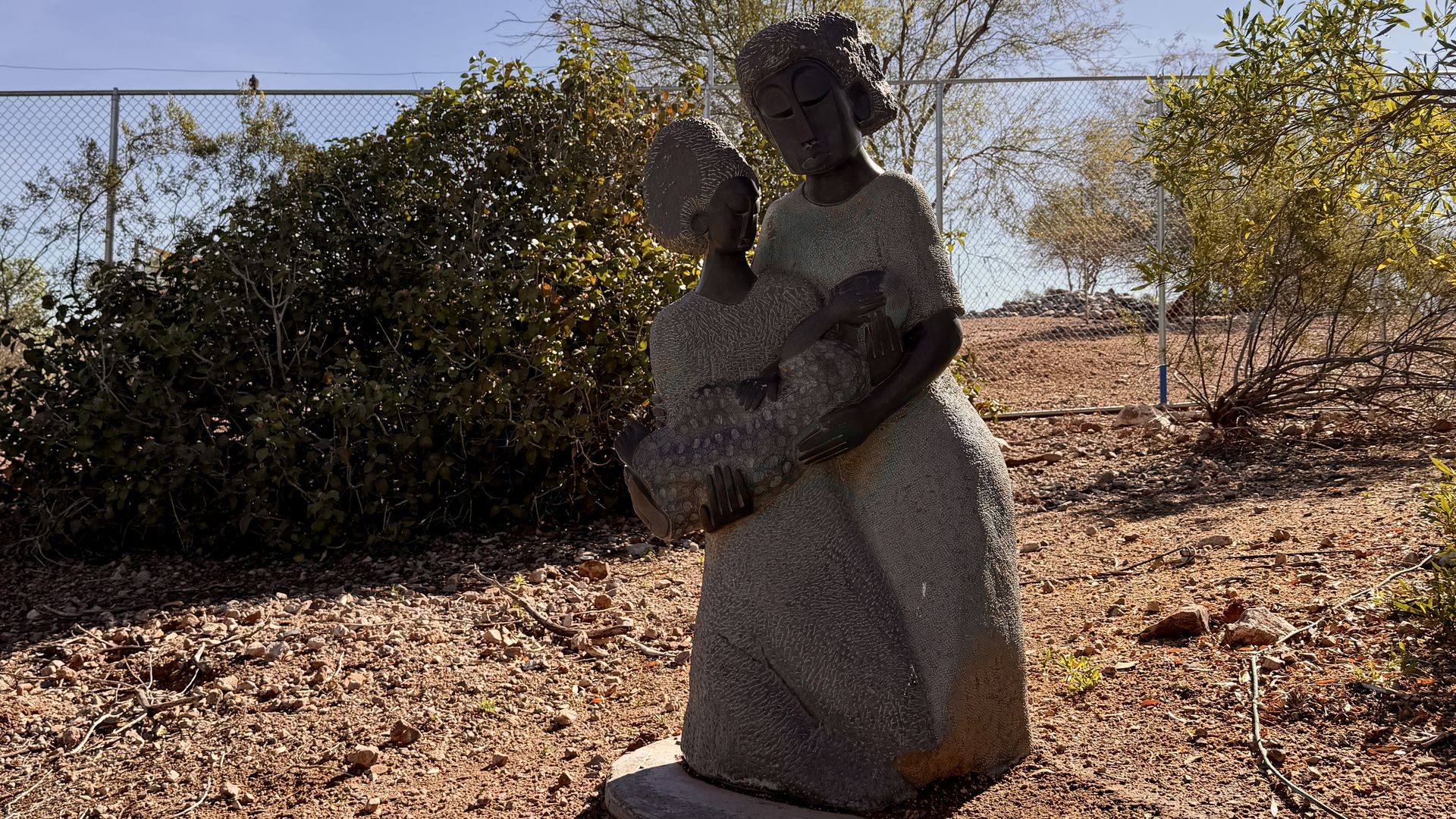 Bronze statue of two women in textured dresses holding a baby, outdoors on dirt with bushes, a fence, and leafless trees under a clear blue sky.