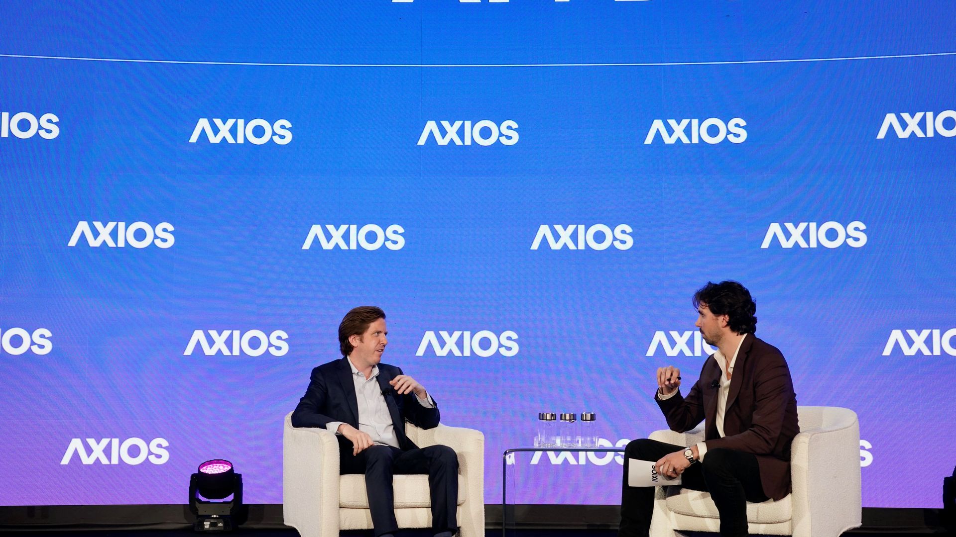 Two men in suits seated on white chairs on stage with blue backdrop displaying AI+DC and Axios logos during a formal discussion or interview event.