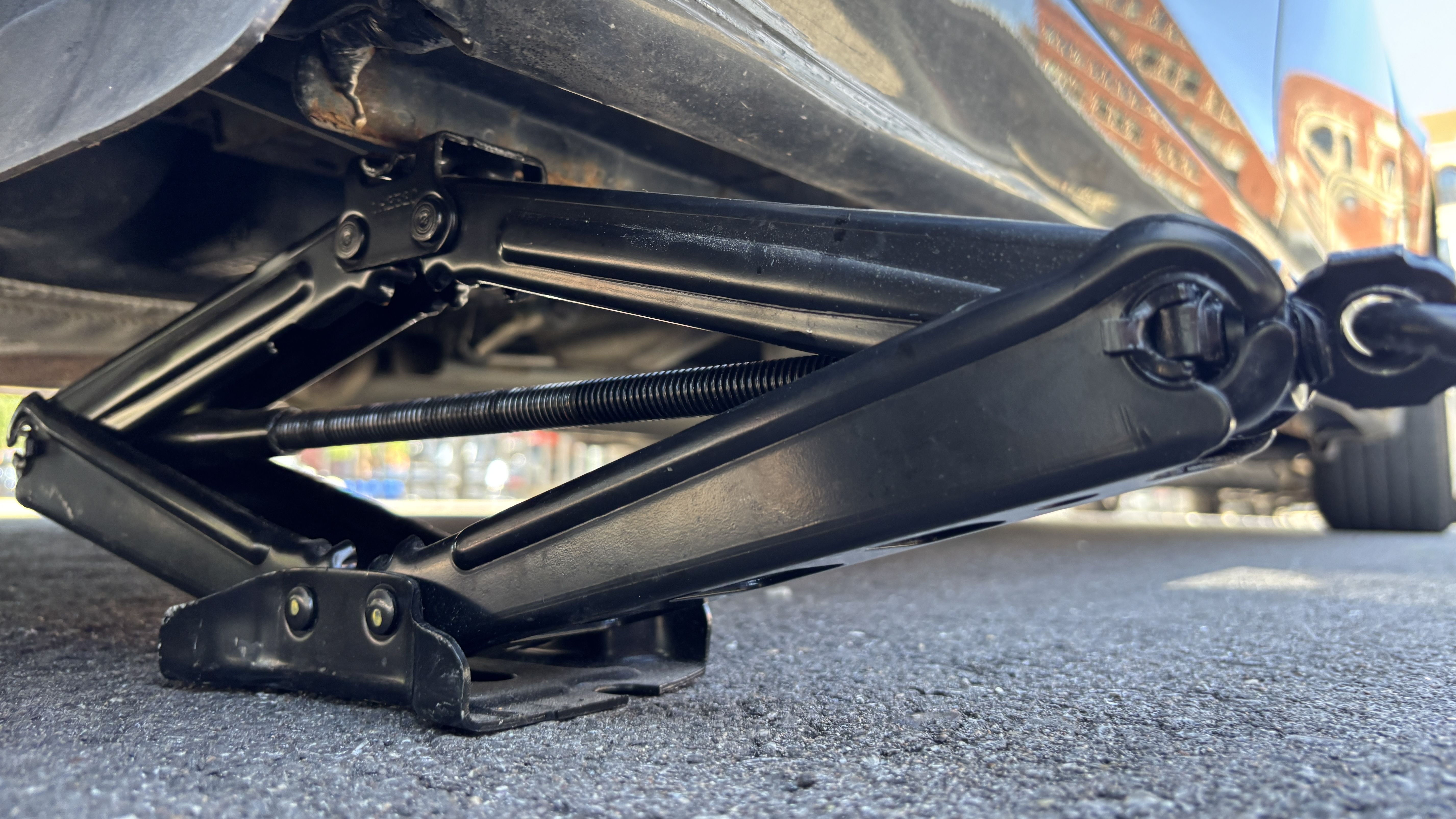 Close-up of a black scissor jack under a car on a paved surface, showing metal arms, bolts, and a threaded rod.