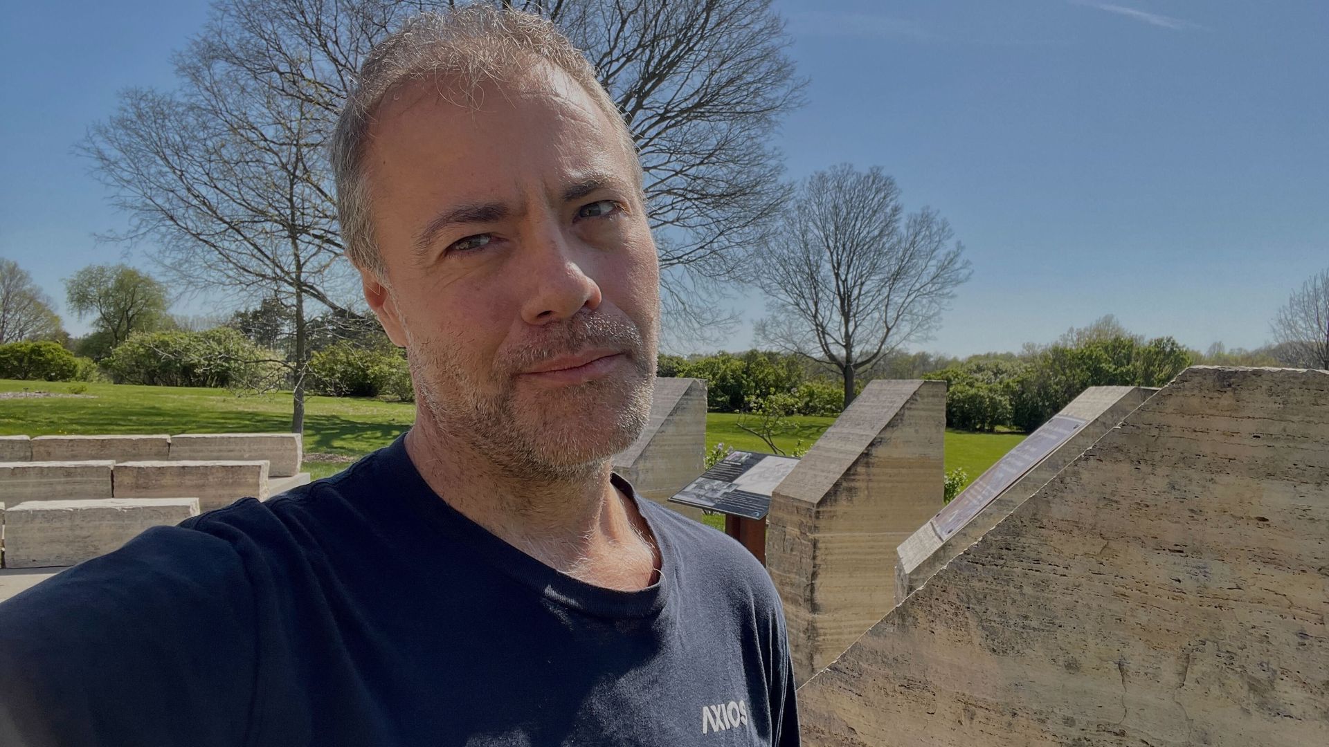 Close-up of a man with gray hair and light stubble taking a selfie outdoors on a sunny day, with leafless trees and large stone steps in the background.