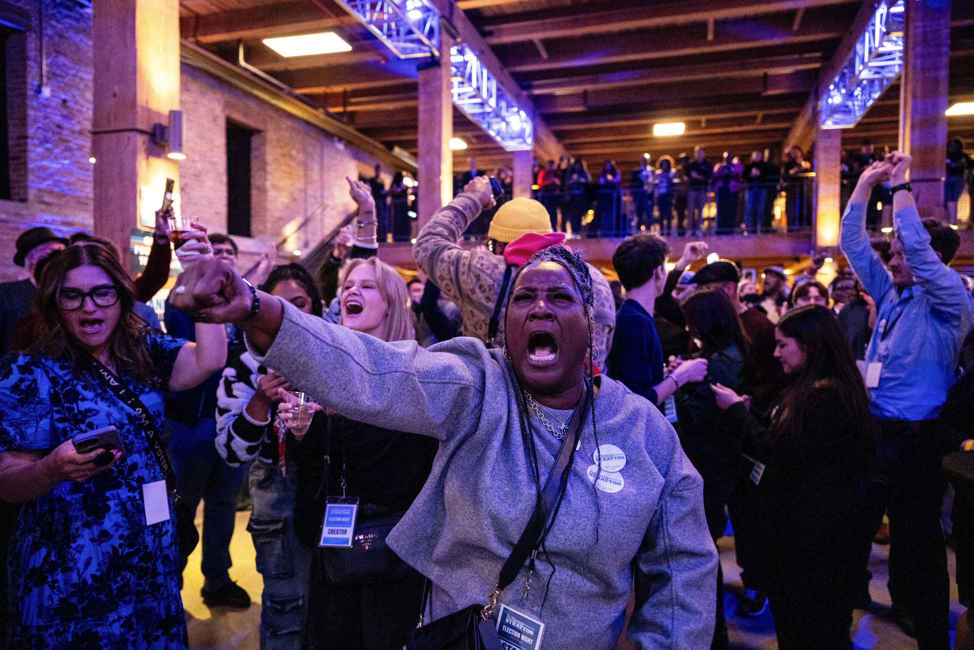 Energetic crowd in a brick-walled venue with blue lighting. A woman in a gray hoodie center-shouts and raises her arm, surrounded by cheering attendees with lanyards and badges.