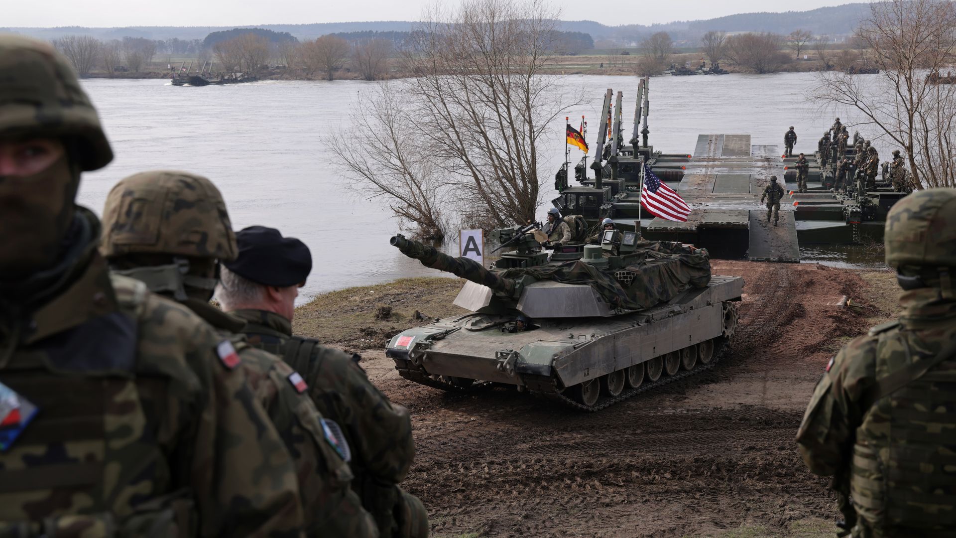 Soldiers in camouflage beside tanks on a floating bridge across a wide river; U.S. and German flags wave as leafless trees and distant hills loom under an overcast sky.