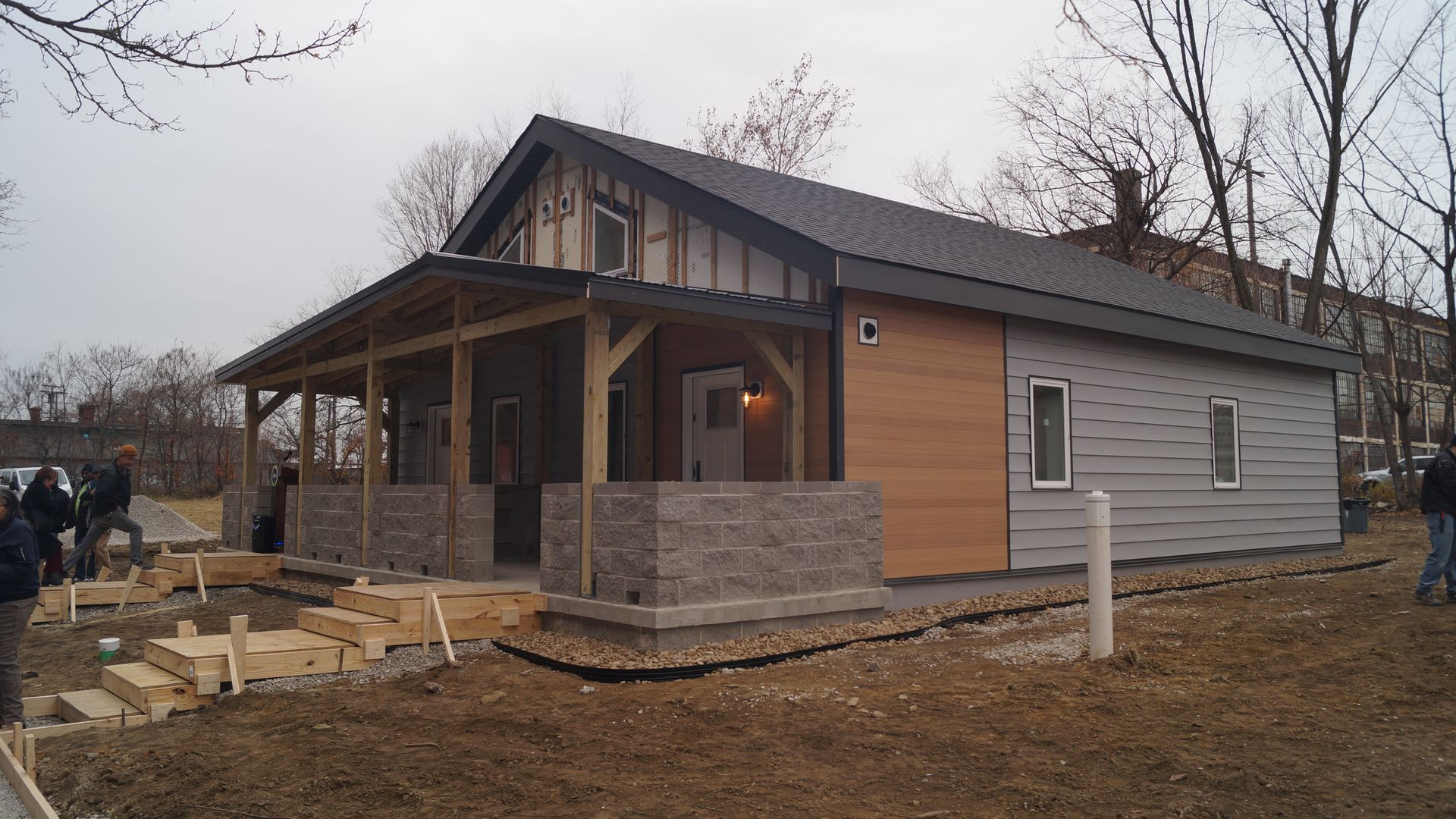 A modern house under construction with gray and wood panel siding, black roof, a small porch with stone half walls, wooden steps, and several people nearby on a cloudy day.