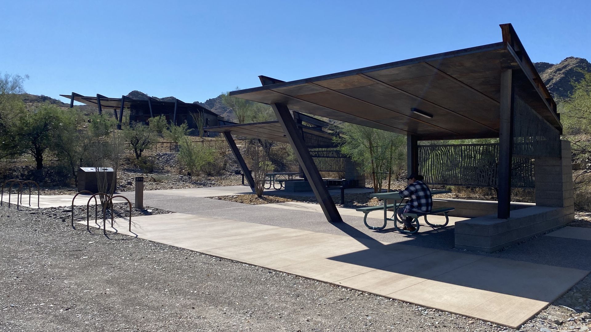 Two metal ramadas in a mountain desert area with a man sitting at bench. 