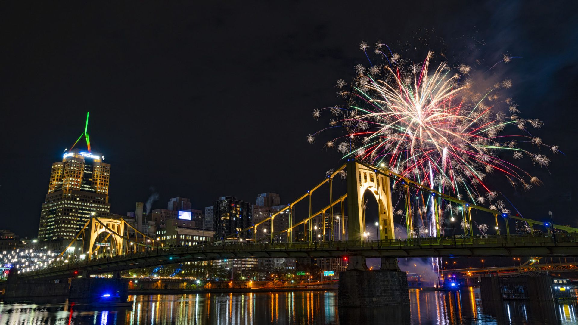 Night view of a yellow suspension bridge with a city skyline in the background, featuring colorful fireworks lighting up the sky above the river.