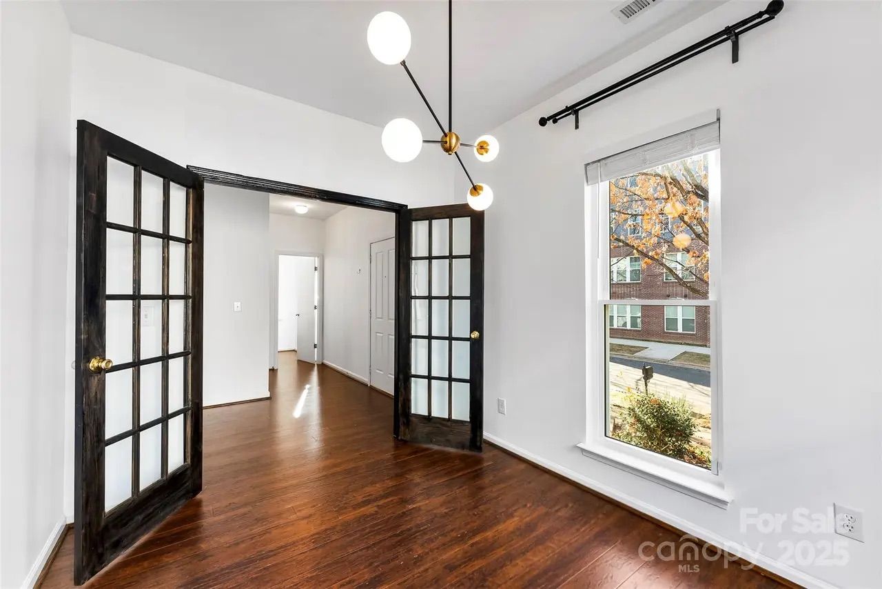 Empty room with dark wooden floor, white walls, large window showing outdoor tree and building, black-framed French doors, and modern ceiling light fixture with round bulbs.