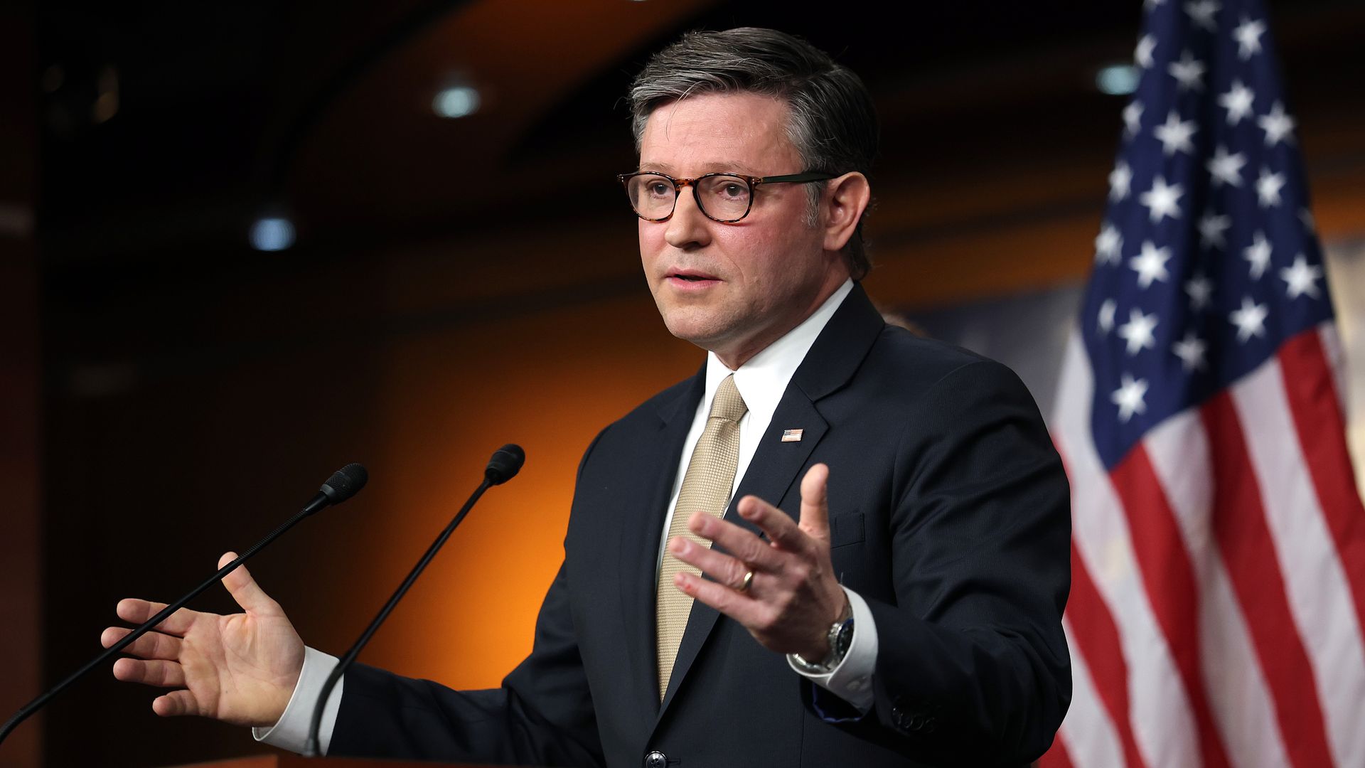 A man in a dark suit and beige tie, wearing round glasses, speaks at a podium with two microphones; an American flag stands behind him, under warm orange lighting.
